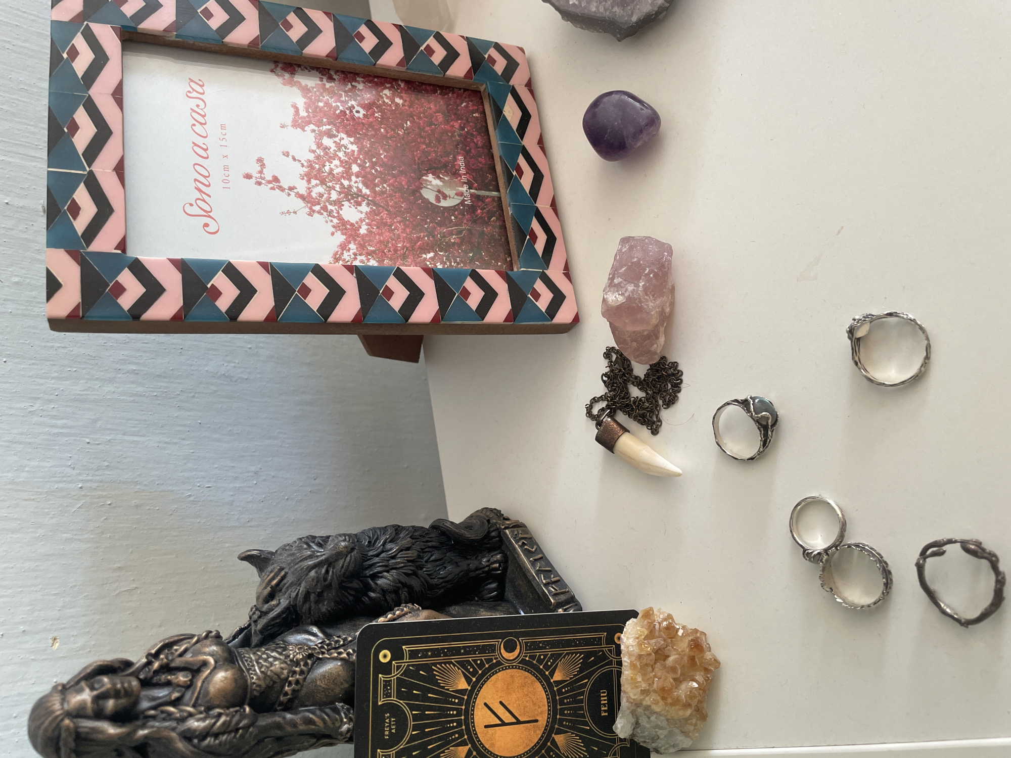 On a white shelf against a pale blue wall, a collection of objects is arranged. To the left, a dark bronze figurine depicts a woman with braided hair and elaborate clothing, seated behind a wolf with its head turned. In front of the figurine, a black card with gold celestial designs and the rune 'Fehu' is visible, along with a cluster of yellow, crystalline stones. To the right of the figurine, a photo frame with a colorful, geometric pattern of pink, teal, and black rhombuses holds a picture of a tree with pink blossoms, and text that reads "Sono a casa" and "Made in India." Further to the right, a smooth, pale pink stone, a small, faceted purple stone, and a larger, rough purple crystal are arranged on the shelf. In the foreground, several rings are scattered across the shelf: two silver bands with intricate designs, a silver ring with a gray stone, and a dark, twig-like ring. A necklace with a light-colored, tooth-shaped pendant and a dark chain is also present.