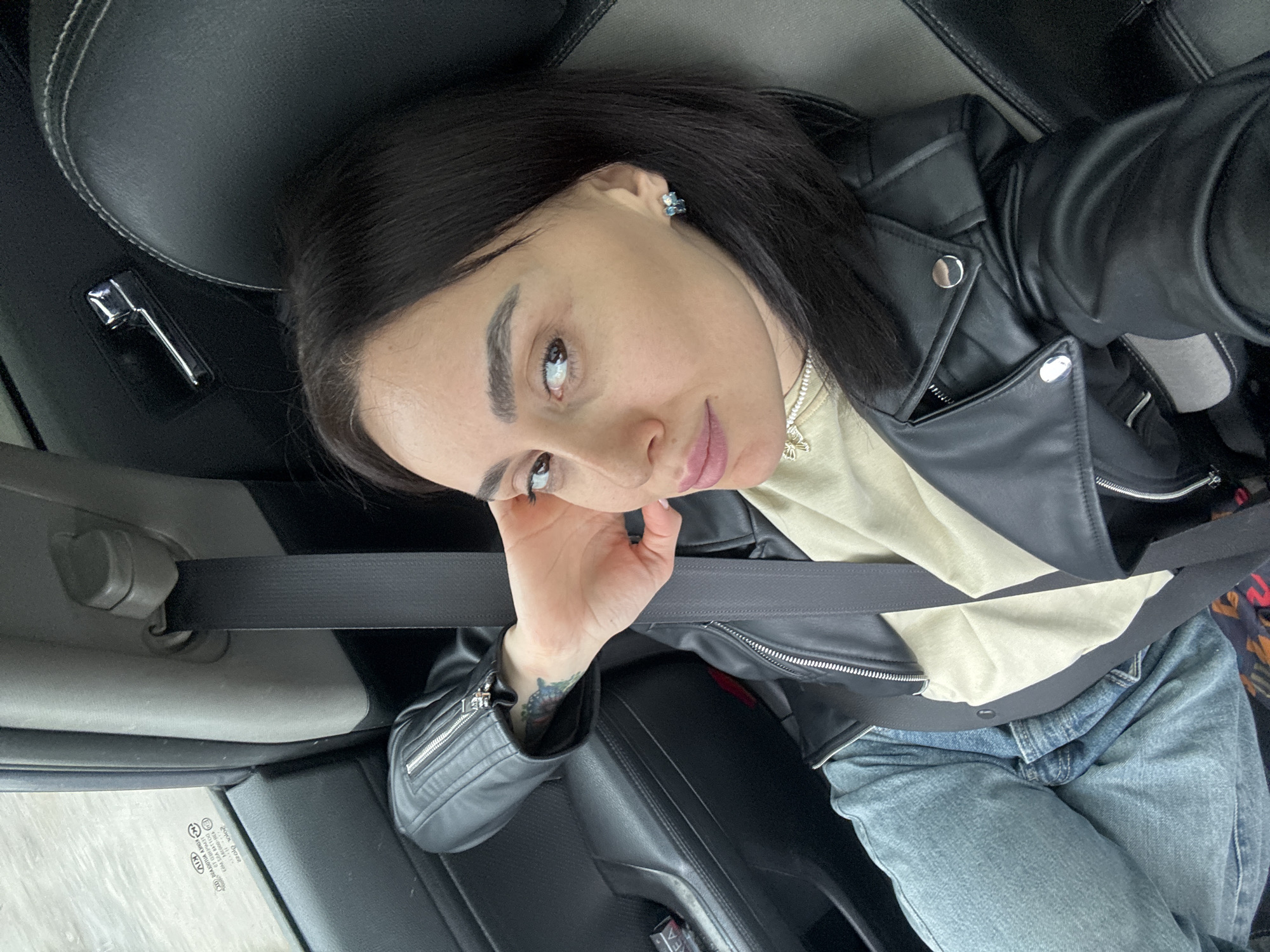 A dark-haired adult female is seated in the passenger seat of a vehicle in Tbilisi, Georgia. She wears a black faux leather jacket over a light beige top and light blue jeans, secured by a dark seatbelt. Her left hand is positioned behind her head, revealing a multi-colored tattoo on her inner forearm. She has a beaded necklace with a gold-toned pendant and small stud earrings. The vehicle interior features black and grey upholstery with white stitching. Visible through the window on the left is a backward-facing sticker displaying "KIA," the Georgian text "თეგეტა მოტორსი" (Tegeta Motors), and a Tbilisi landline phone number starting with "032," confirming the location. A small black zippered pouch rests on the armrest on the left.