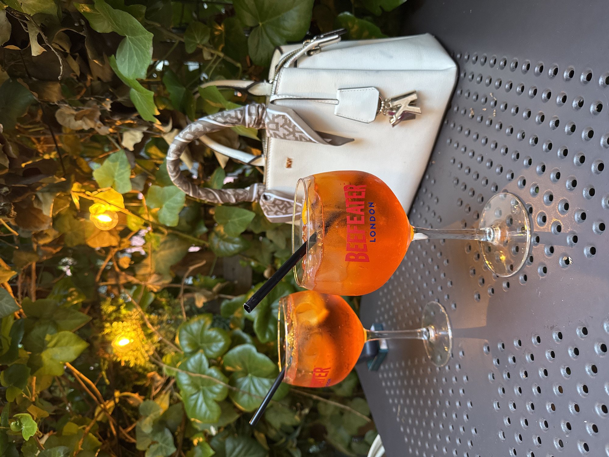 An outdoor, nighttime scene depicts two stemmed glasses filled with an amber-orange cocktail, containing ice cubes and citrus slices, on a dark grey perforated metal table. Both glasses feature 'BEEFEATER LONDON' in red lettering and contain black drinking straws. To the right, a white DKNY handbag with a patterned brown and white scarf tied around its handle rests on the table. A white leather tag with a silver-colored 'K' charm hangs from the bag. The background consists of lush green foliage, illuminated by numerous warm yellow string lights, indicating an evening setting in the city of Unknown, Unknown.