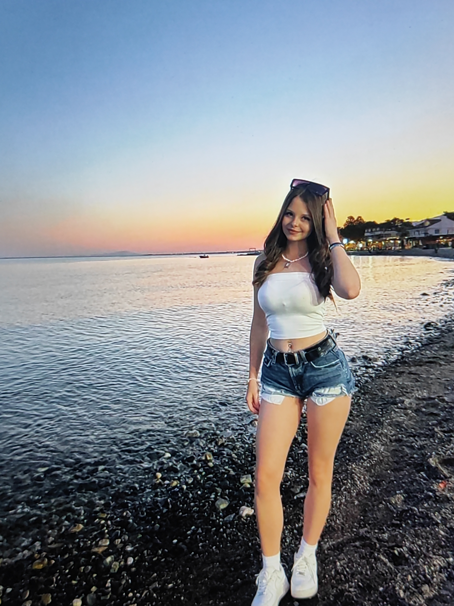 A young woman stands on a pebble beach, facing forward and smiling faintly. She is wearing a white strapless top, denim shorts with white trim, a black belt with a silver buckle, and white sneakers. She has long, wavy brown hair and is wearing a necklace and sunglasses pushed up on her head. The water of a sea or large lake is to her left, with gentle waves. In the background, across the water, is a coastline with buildings and trees, illuminated by the warm light of a sunset. The sky displays a gradient of blue fading into orange and yellow. The location is not the city of Fergana, Uzbekistan; it appears to be a coastal area.