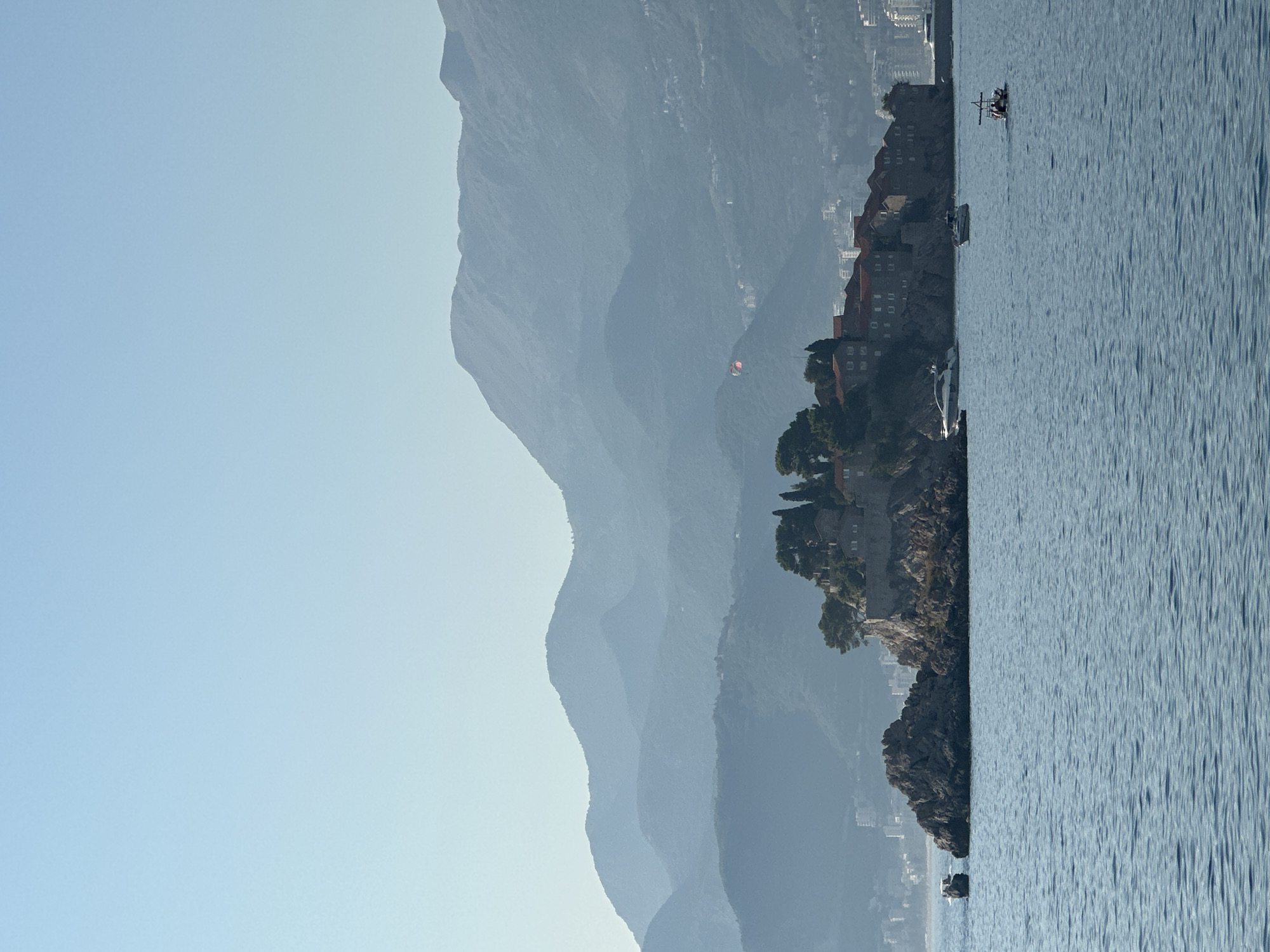 The image depicts the island of Sveti Stefan in Montenegro, with its historic stone buildings and red-tiled roofs situated on a rocky outcrop. The island is connected to the mainland by a narrow causeway. The calm, blue waters of the Adriatic Sea are in the foreground, with several boats visible: a small speed boat with two people, a larger white yacht, and another smaller vessel. In the background, hazy blue mountains form a layered silhouette against a pale sky. The overall atmosphere is serene and picturesque, suggesting a clear, warm day. There is a paraglider visible in the far distance, over the mountains.