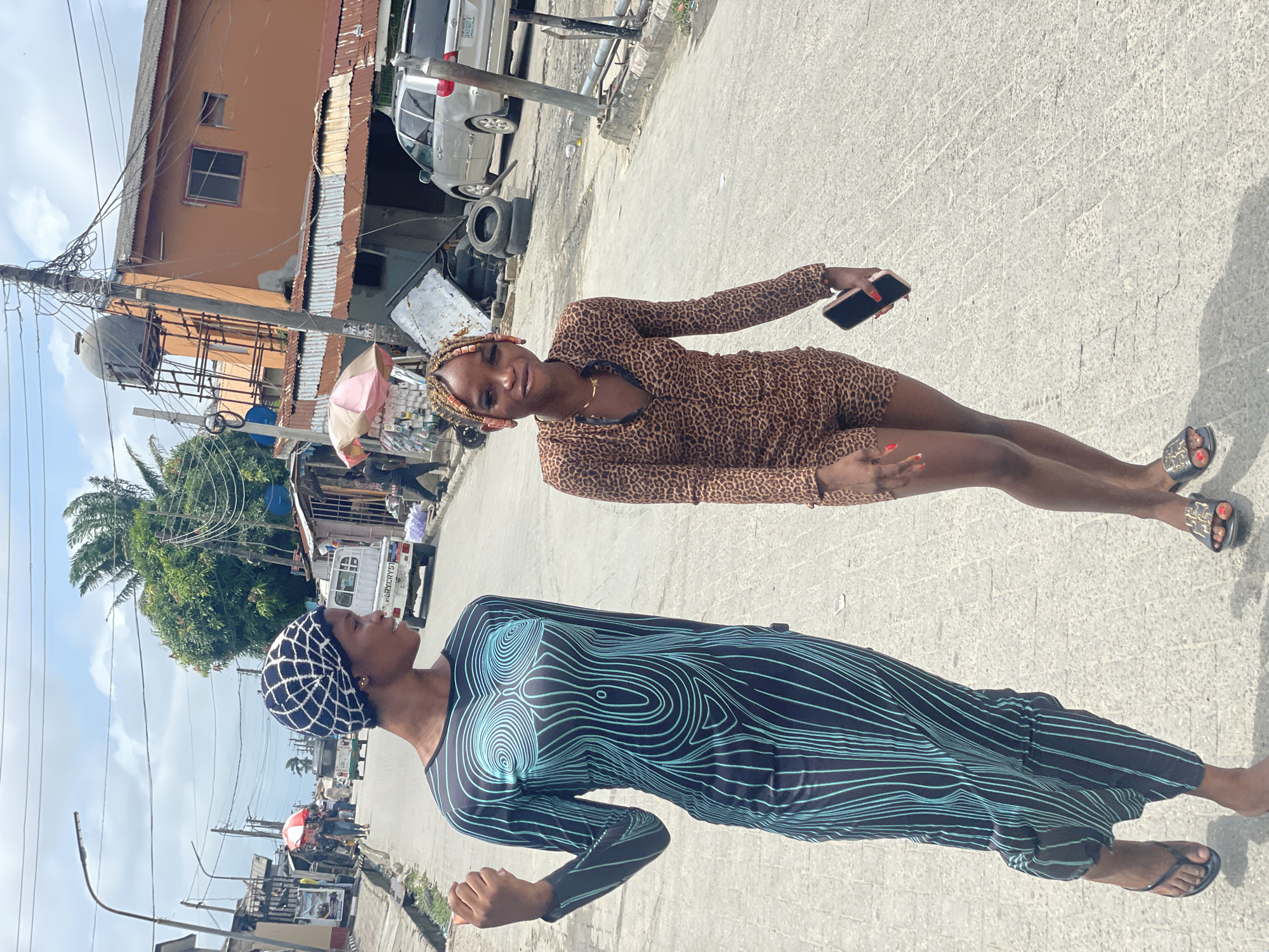 The image depicts two women standing on a concrete or paved street in what appears to be a commercial or residential area, likely in the city of Ado, Nigeria based on the environmental cues. The woman in the foreground is wearing a long-sleeved dress with a blue and black swirl pattern. She also wears a dark patterned head covering. The other woman in the background wears a short, long-sleeved leopard-print dress and holds a cell phone in her hand. There are buildings, utility poles, and parked vehicles visible in the background, suggesting an urban setting. The sky is partly cloudy. The street has visible cracks and texture. The image is oriented in a landscape format.