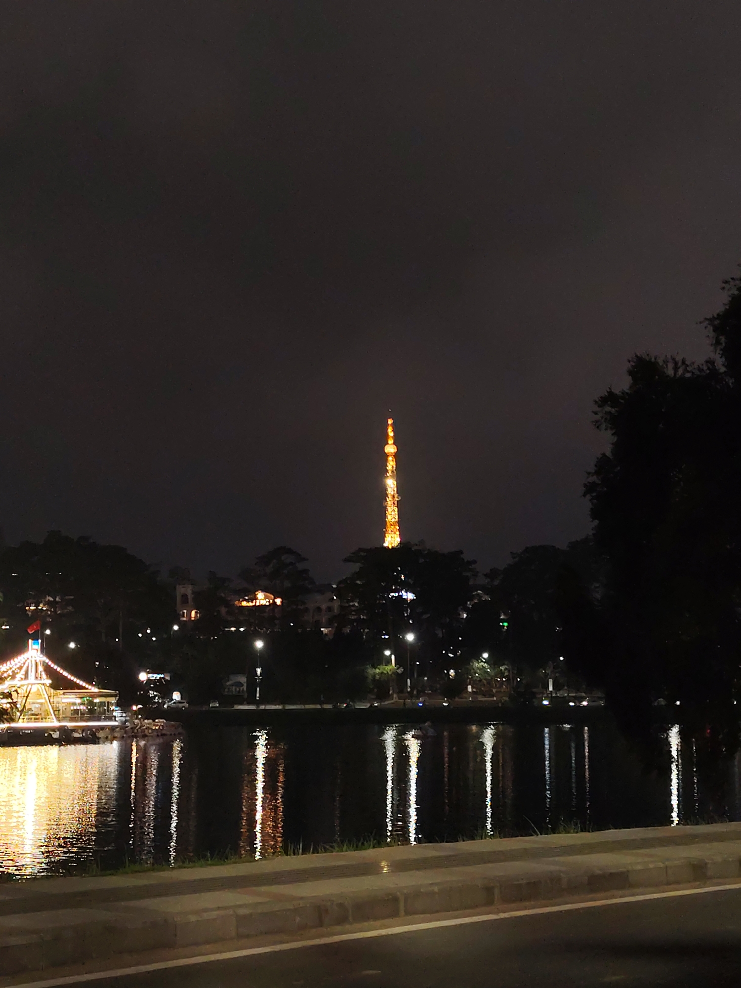 A nighttime view in Da Lat, Vietnam, shows a body of water reflecting lights from buildings and the Eiffel Tower-like structure in the background. A building, possibly a restaurant or attraction, is lit up on the left side of the water. Trees outline the skyline. The foreground includes a walkway or road.