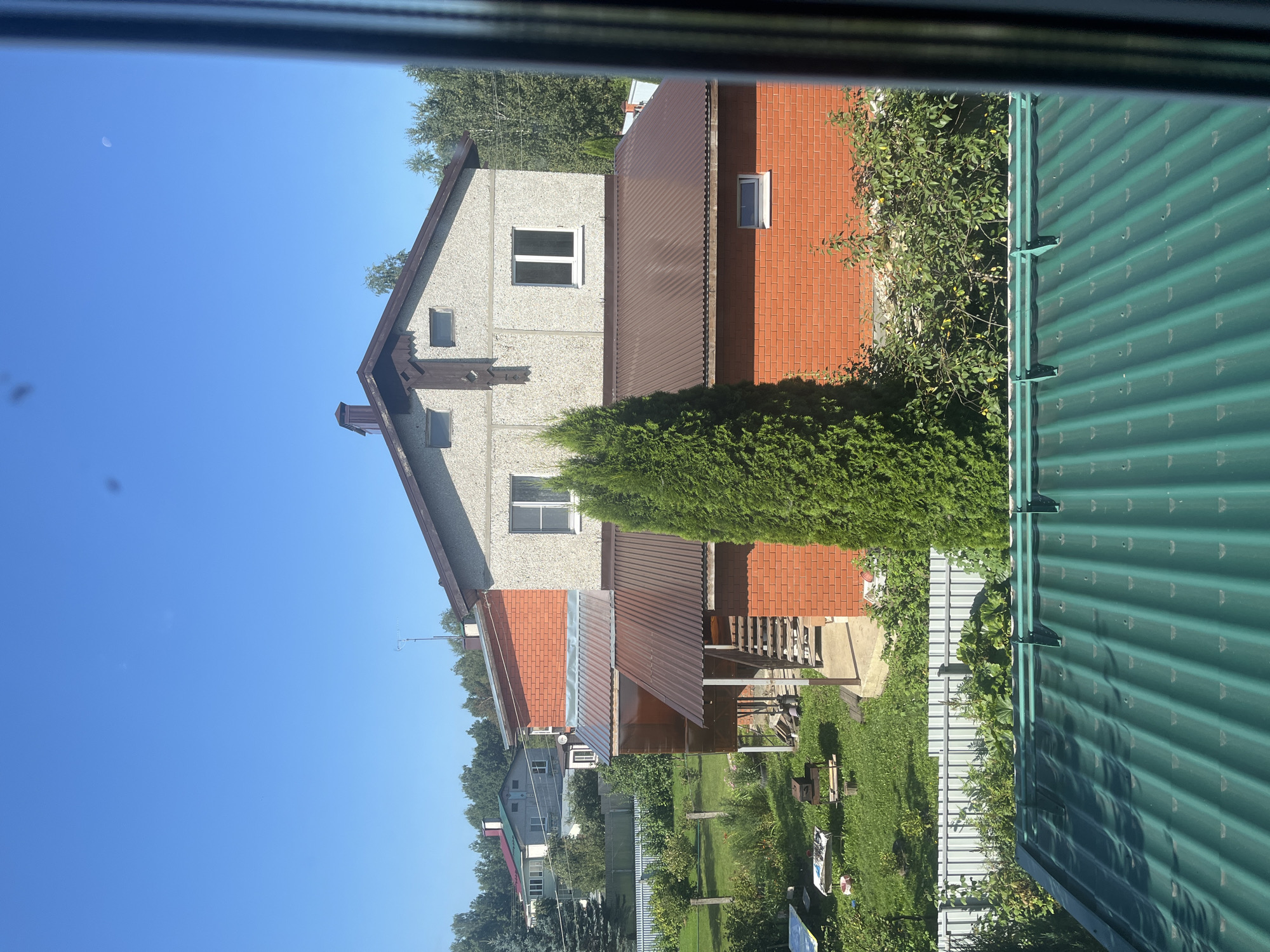 A view of residential houses and surrounding vegetation under a clear blue sky. The foreground features a green corrugated metal roof, likely belonging to an outbuilding or garage. Behind this, a two-story house with a brick lower section and a textured grey upper section is visible. This house has a brown metal roof with a chimney and multiple windows. Adjacent to it is another section of the same house, clad in red brick, with a single window. A covered porch area with a metal roof extends from this brick section, leading to an outdoor staircase. Further in the background, a long, light-colored fence separates the property from more houses and lush green trees. The overall setting appears to be a suburban or rural area, consistent with a location like Novy, Russia, characterized by private residences and abundant greenery. The image captures a daytime scene, with bright sunlight casting shadows. No people or animals are present in the frame.