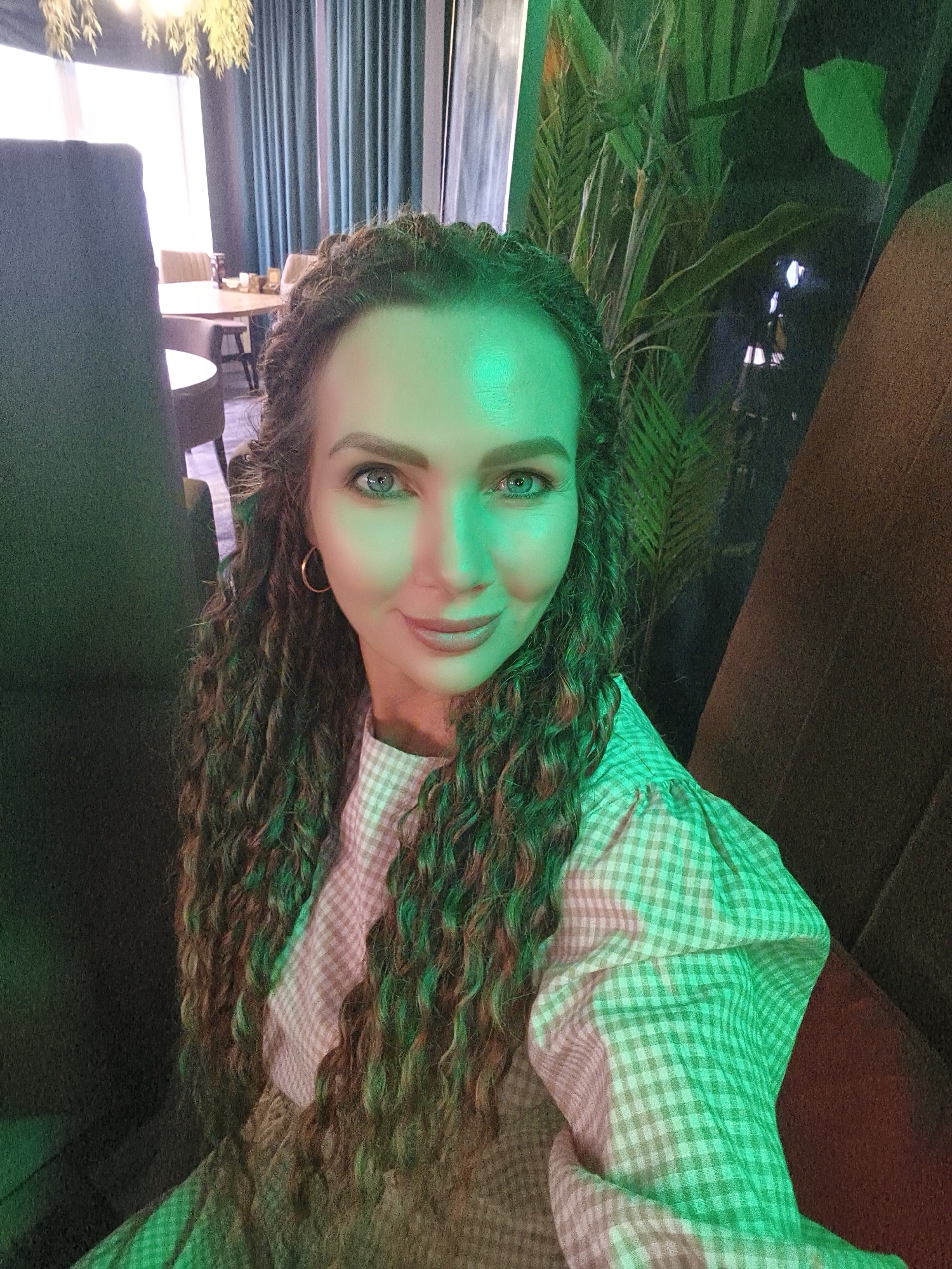 The image shows a woman with long, curly, brown hair with blonde highlights, wearing a light-colored checkered shirt. She is looking at the camera with a slight smile and her right eye is partially obscured by green lighting. The lighting in the scene is predominantly green, casting a strong hue on her face and hair. The background is dimly lit and appears to be an indoor setting with dark furniture, green foliage from plants, and a glimpse of tables and chairs, suggesting a restaurant or lounge area. The woman is seated, with her body angled towards the right side of the frame.