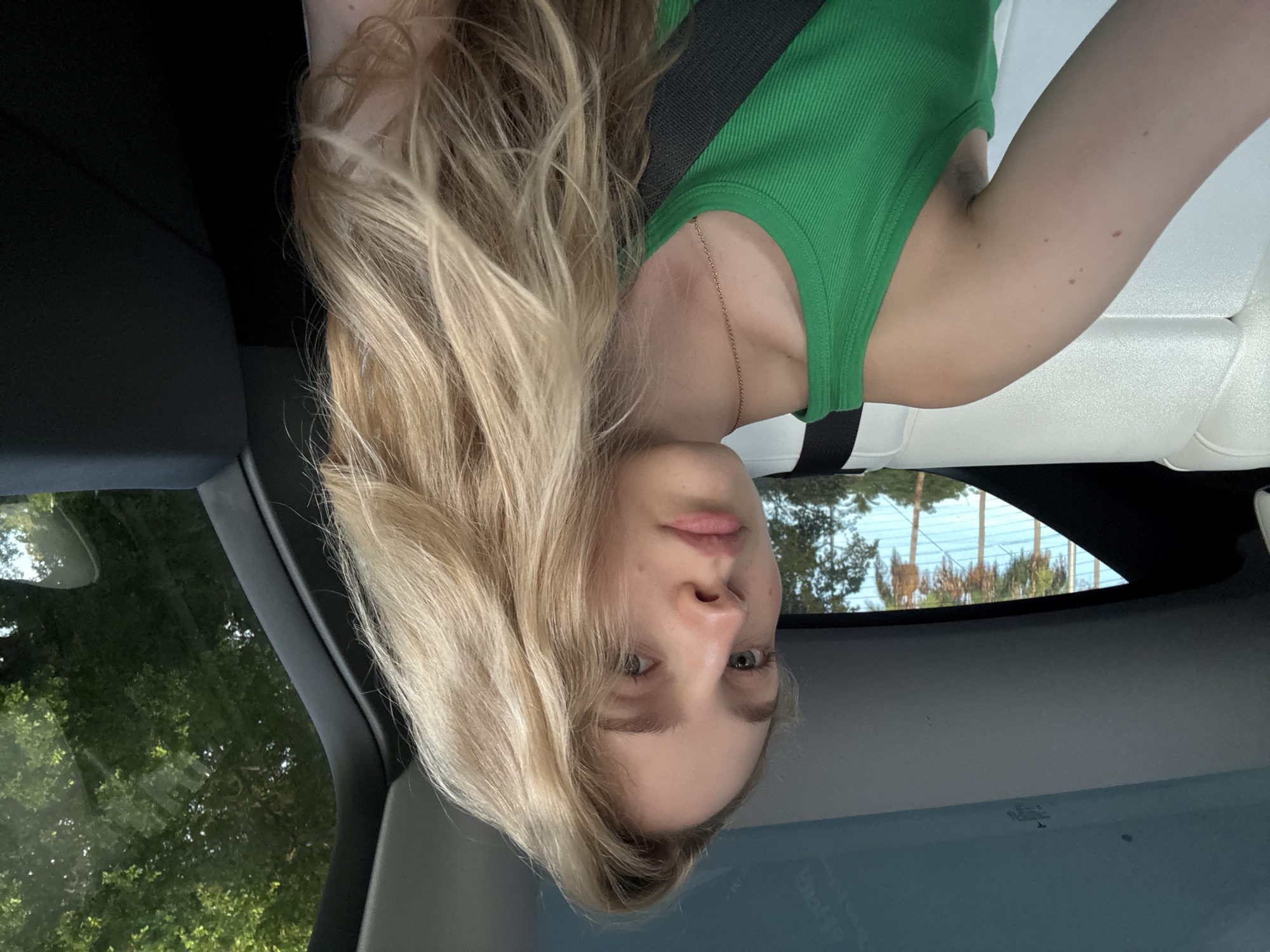 A young woman with blonde hair is seated in a car, leaning back. She is wearing a green tank top and a necklace. Her face is turned towards the camera, and she has light blue eyes and light-colored eyebrows. Her hair is long and wavy, falling around her face and shoulders. The interior of the car appears to be white leather. Through the car's windows, trees and what appear to be palm trees are visible, suggesting an outdoor setting. The lighting suggests it is daytime. The image appears to be a selfie taken from inside the car. The background outside the car is bright, indicating a sunny day. There is no visible text in the image.