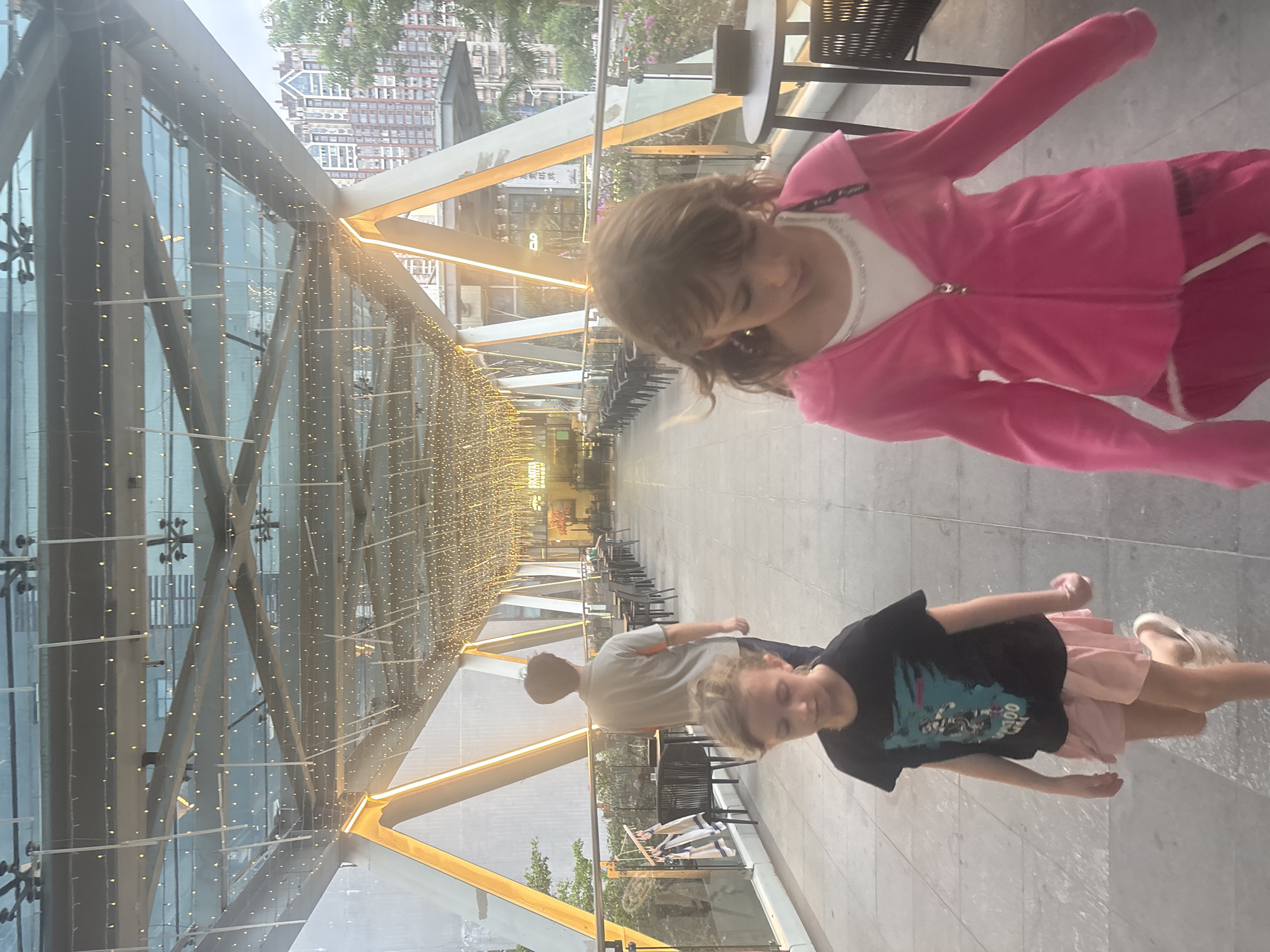 In the Liwan District of China, two young girls and a man are captured in a long, covered walkway. The walkway is illuminated by strings of small lights and has a glass ceiling and sides, with prominent orange-yellow triangular support beams. In the foreground, a girl in a black t-shirt and pink shorts walks towards the viewer, while another girl in a pink hooded jacket and pink pants walks ahead. A man, seen from behind, walks away from the viewer further down the walkway, which is lined with tables and chairs, suggesting an outdoor dining area. Beyond the walkway, modern buildings are visible. The overall scene depicts a pedestrian area within a city.