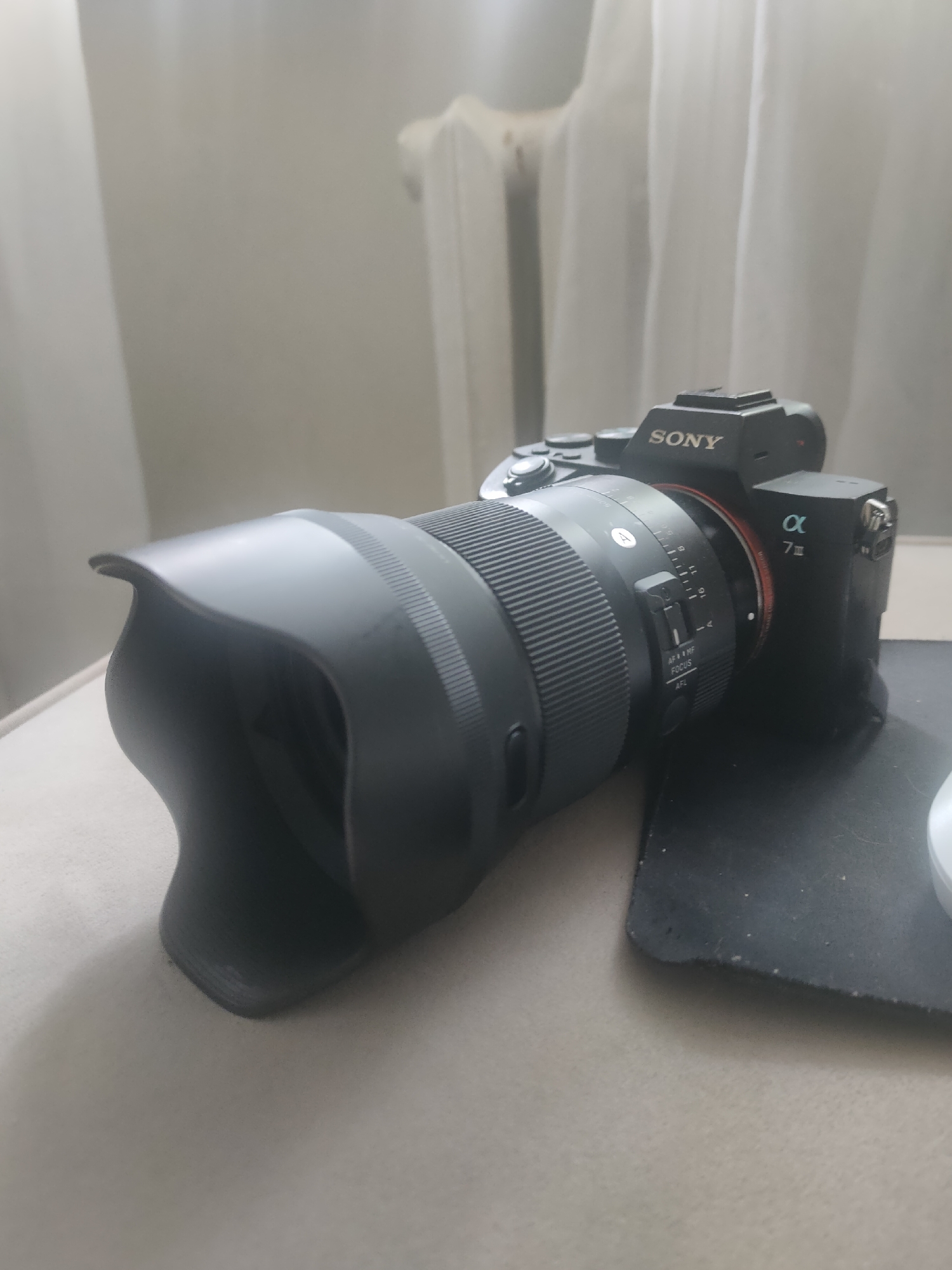The image shows a black Sony Alpha 7 III camera with a Sigma lens attached, resting on a textured surface, likely a table or a soft fabric. The camera has the "SONY" logo visible on the top and the "α 7III" designation on the back. The lens has a prominent lens hood. The background consists of white curtains or blinds, suggesting an indoor setting. There are no people visible in the image. The lighting appears to be natural and diffused, possibly from a window. The location is not specified in the image.