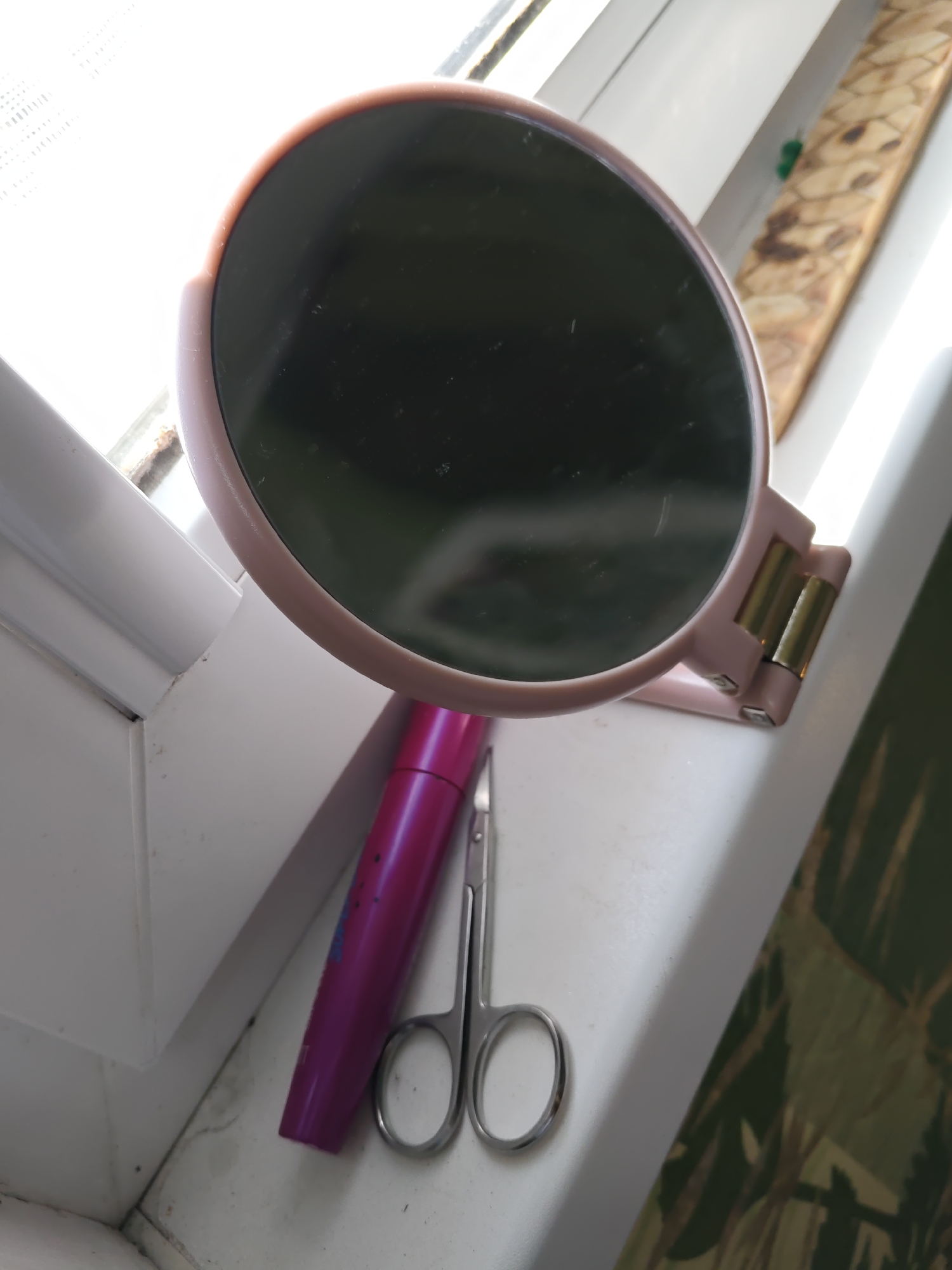 A close-up, eye-level shot shows a pink hand mirror with a gold hinge resting on a white windowsill. To the left of the mirror is a purple mascara wand. To the right of the mascara wand are small, silver scissors. The background of the image is a window with a view of green foliage outside. The scene is located on a windowsill in Rostov-on-Don, Russia.