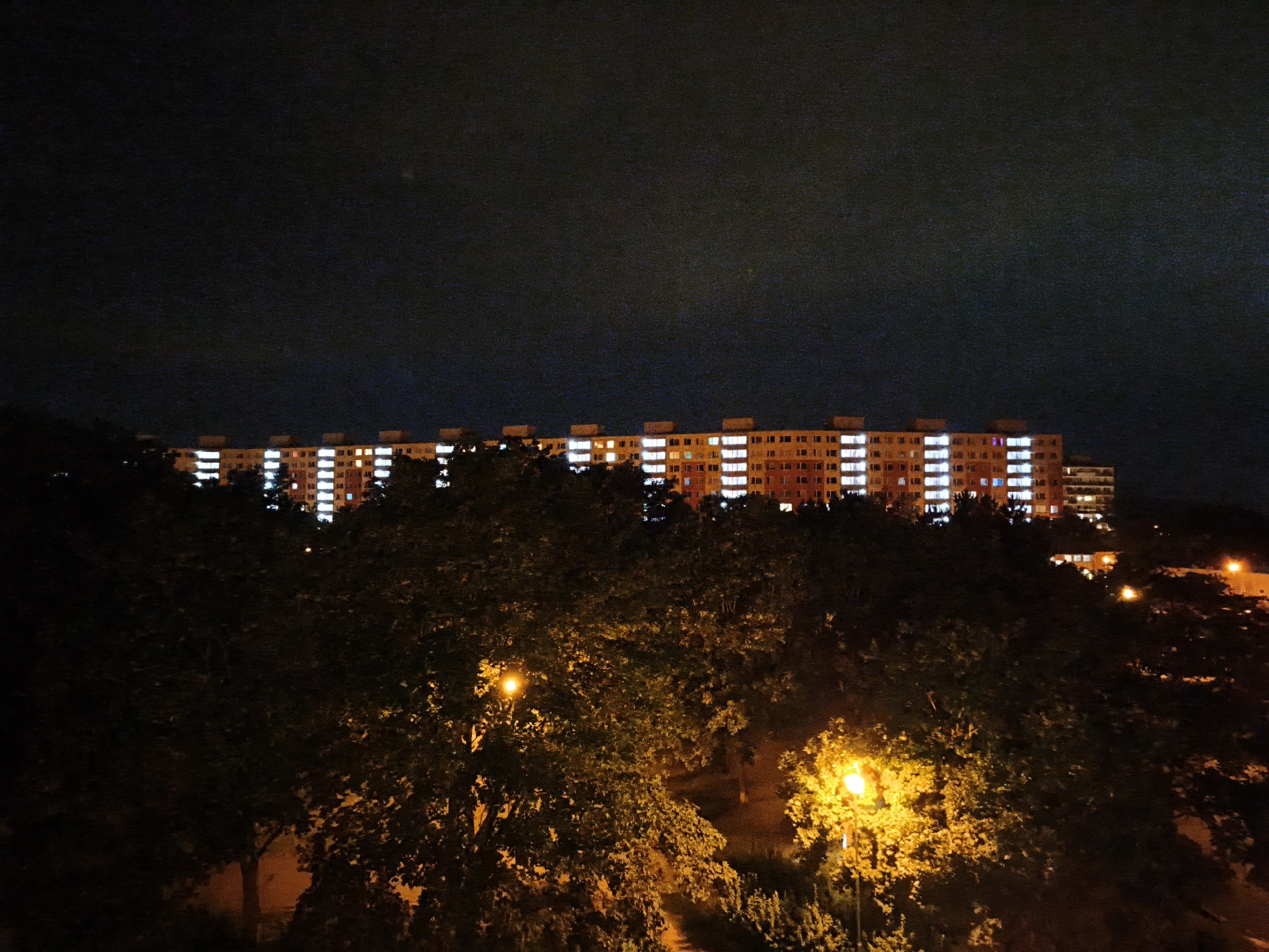 The image depicts a long, multi-story apartment building at night, with many windows illuminated. The building is located in the city of Most, Czechia. In the foreground, silhouetted trees and streetlights create a dark, textured layer between the viewer and the building. The sky above is dark and appears to be clear.