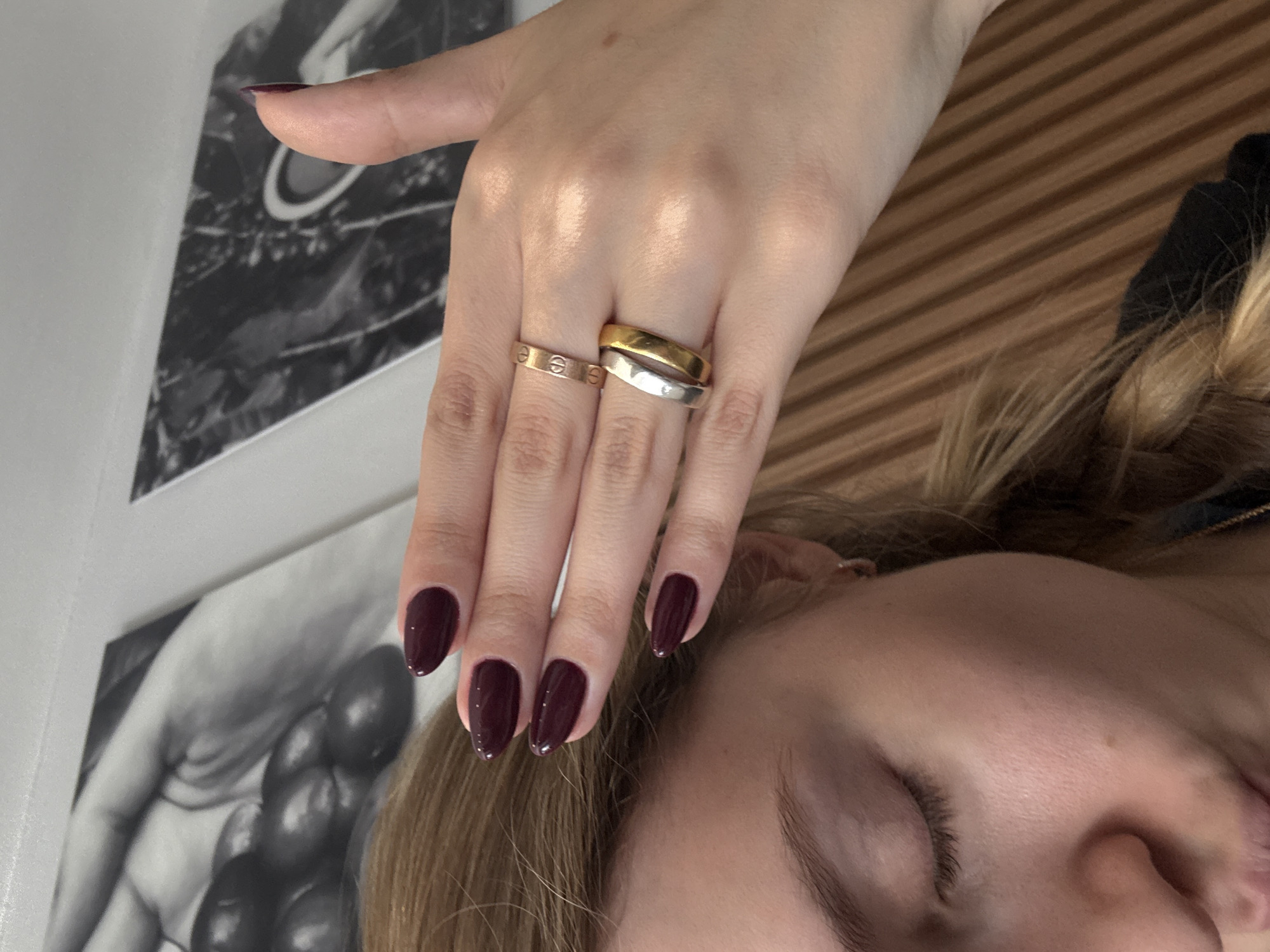 A person is seen from an overhead perspective with their eyes closed, a section of blonde hair visible at the lower right. The person's left hand, adorned with two rings on one finger and one on another, is extended towards the upper left. The nails are manicured with a deep burgundy color. The background shows a black and white print and horizontal wooden surface. The location is unknown.