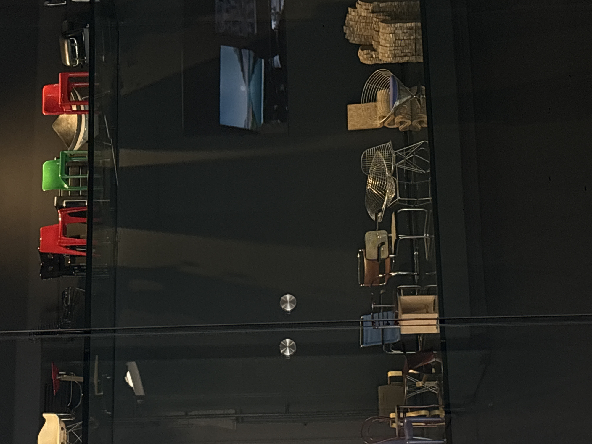 The image shows a display of various chairs behind a glass wall in Mexico City, Mexico. The chairs are stacked and arranged in a vertical line, showcasing a variety of styles and colors, including red, green, and beige. The glass wall is divided into sections, held together by metal hardware. The background is dark.