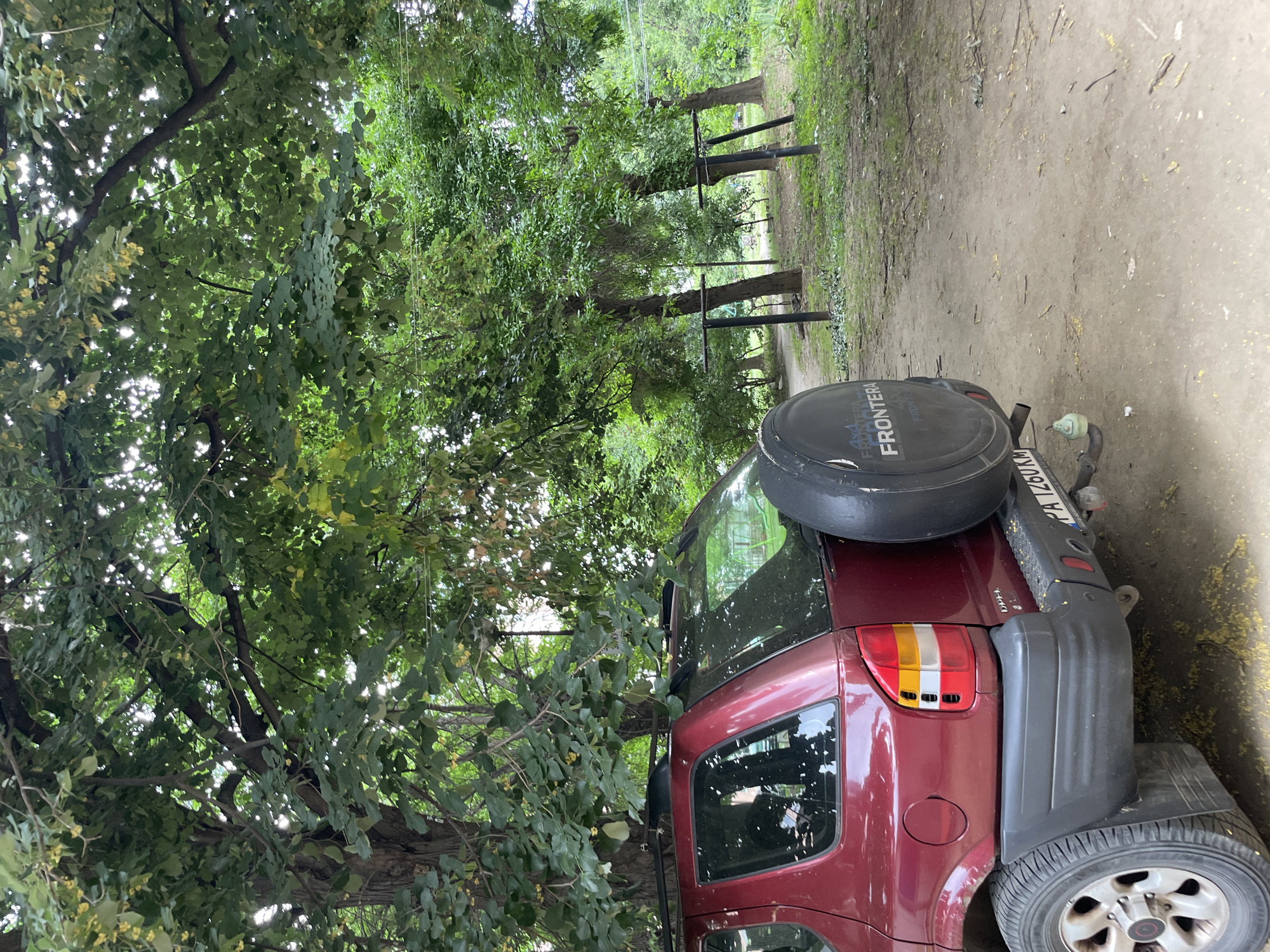 A dark red SUV is parked on a dirt road lined with trees. The vehicle has a spare tire mounted on the back. The trees have lush green foliage, and the sun is shining through the leaves, creating dappled light. The scene appears to be in a rural or wooded area. There is some text visible on the spare tire, which reads "FRONTERA." There is also a license plate visible on the back of the vehicle, but the text is not legible. The overall mood of the image is peaceful and natural.