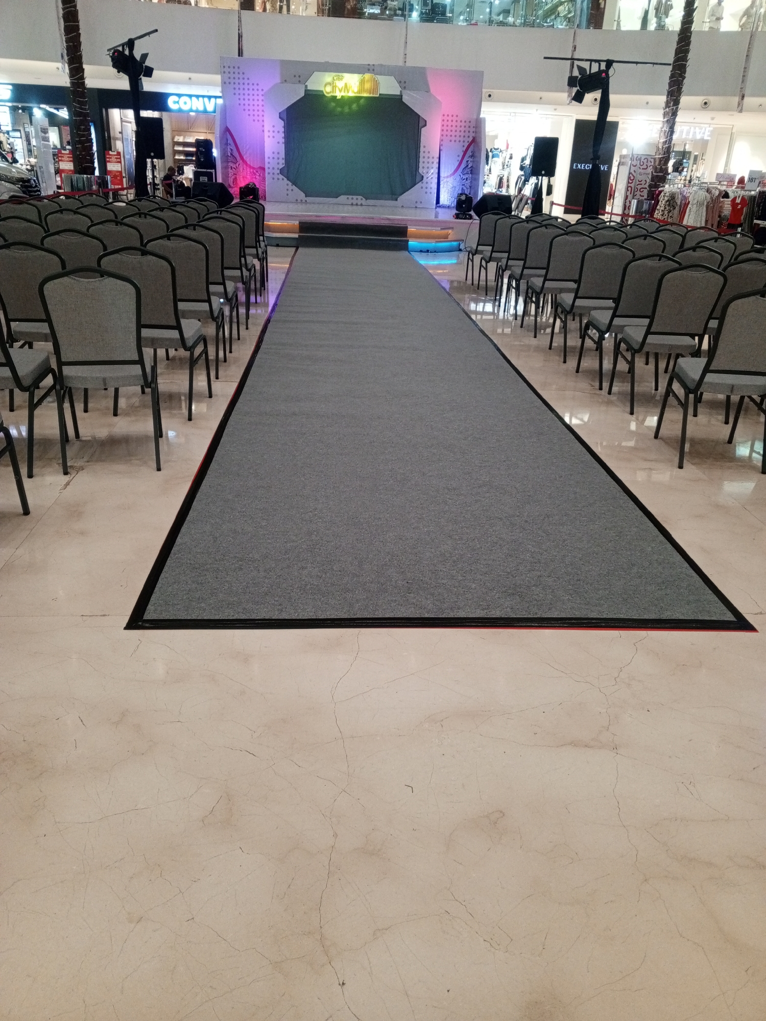 The image depicts a runway set up inside a shopping mall, likely for a fashion event or product launch. A grey carpeted runway extends from the foreground towards a stage at the back of the mall. Rows of empty grey chairs are neatly arranged on either side of the runway. The stage features a large backdrop with lighting and a logo that appears to say "GIBR CityMall," indicating the location is likely Cibinong City Mall in Cibinong, Indonesia. Various retail store fronts are visible in the background, suggesting the event is taking place in a public commercial area. The floor is tiled with a light-colored marble pattern. No people are actively engaged in any activity, giving the scene a sense of preparation or a pause in the event.