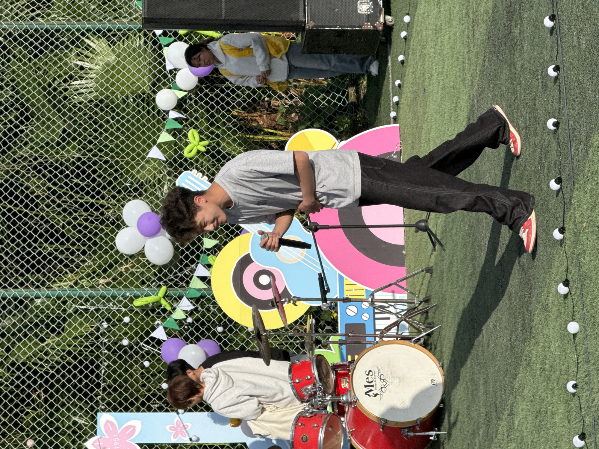 An outdoor musical performance is underway on artificial green turf, likely during a sunny day given the bright lighting and visible shadows. The scene is adorned with colorful decorations, including white and purple balloons, green and white triangular bunting strung along a chain-link fence, and a green balloon animal. Behind the performers, a vibrant backdrop features abstract shapes in blue, pink, yellow, and black, with a stylized guitar motif.
In the foreground, a young man with dark, curly hair, wearing a light grey t-shirt, dark jeans, and red and white sneakers, stands holding a microphone. He appears to be singing or addressing an audience. To his left, partially obscured by a red drum set, another person with short brown hair, wearing a light-colored hoodie, is leaning over the drums, actively playing. The bass drum prominently displays "Mes" and "Drums in Europe."
In the background, behind the chain-link fence and some greenery, a third person with long dark hair, dressed in a grey hoodie with a yellow accent and blue jeans, observes the performance. Two dark speaker or amplifier units are positioned near the fence. String lights with white bulbs are laid out on the turf, adding to the festive atmosphere. A light blue sign with cherry blossom designs and the number "1951" is partially visible in the bottom left corner. The overall setting suggests a casual, outdoor event or concert.