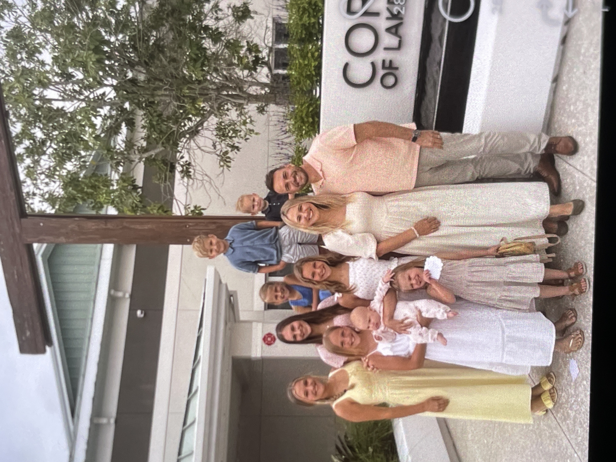 In an outdoor scene, a family of ten, comprising two adults and eight children, poses for a photograph. The two adults, a man and a woman, stand in the foreground on the right side of the image. The man, wearing a pink polo shirt and tan pants, is smiling. The woman, in a cream-colored dress, also smiles. Behind them, to the left, a group of six girls and two boys are arranged. The children are dressed in various outfits, including dresses and shorts. One of the girls is holding a baby. The family is standing on a paved surface in front of a modern building with a horizontal dark band and large white lettering that reads "COR OF LAK28". A large wooden cross structure is visible behind the group. Green trees and shrubbery are also present in the background. The sky is overcast.