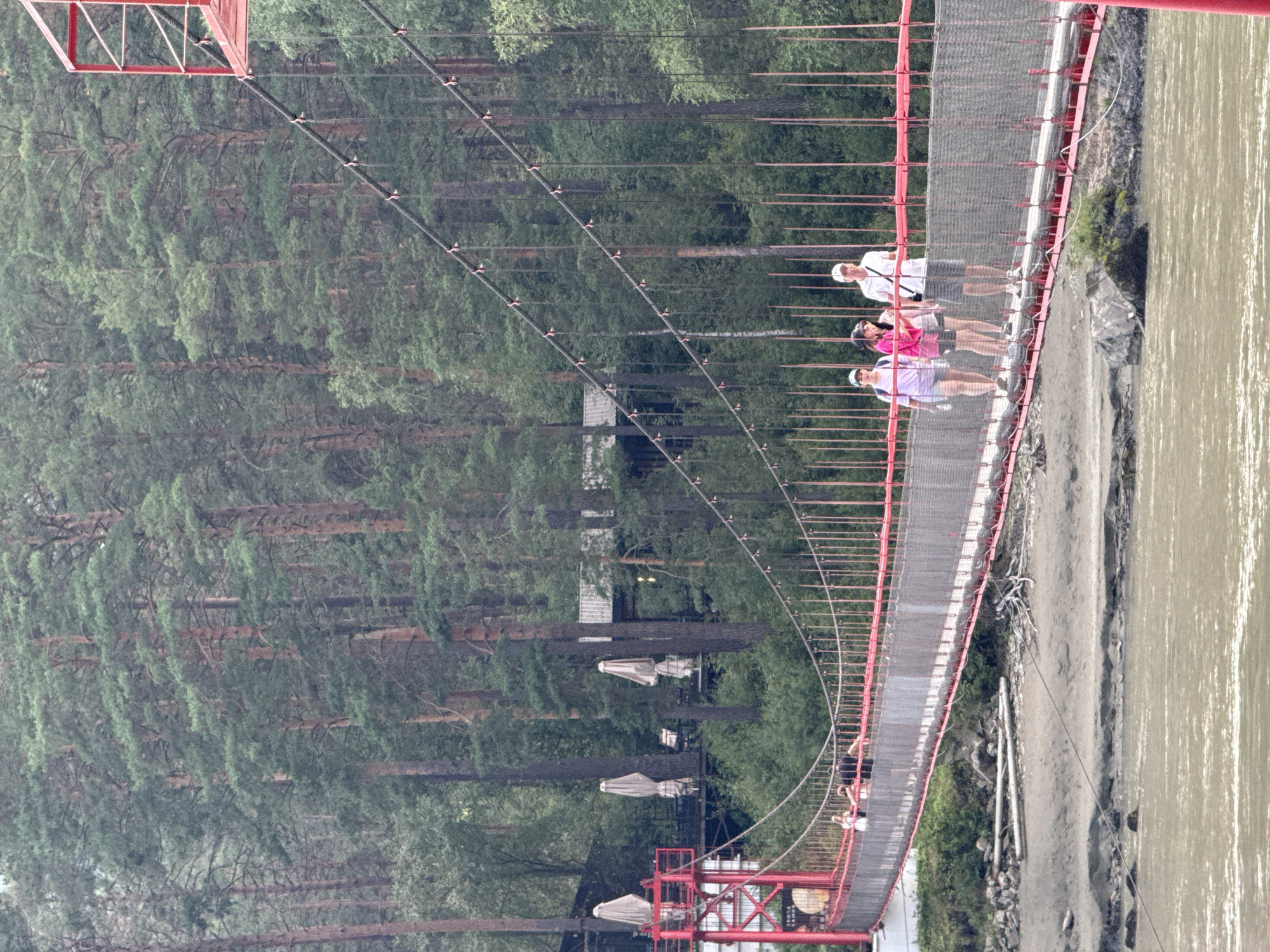 In the city of Gorny Altai, Russia, a long, red suspension bridge spans a wide, murky river. The bridge, constructed with metal cables and a mesh railing, carries four people walking across its surface. The backdrop is a dense forest of tall, green pine trees. On the far bank of the river, nestled within the trees, are buildings with white umbrellas, suggesting a resort or restaurant. The bridge's red structure is visible on the left side of the image, with its red supports and walkways. The sky is overcast, contributing to the diffused lighting of the scene.