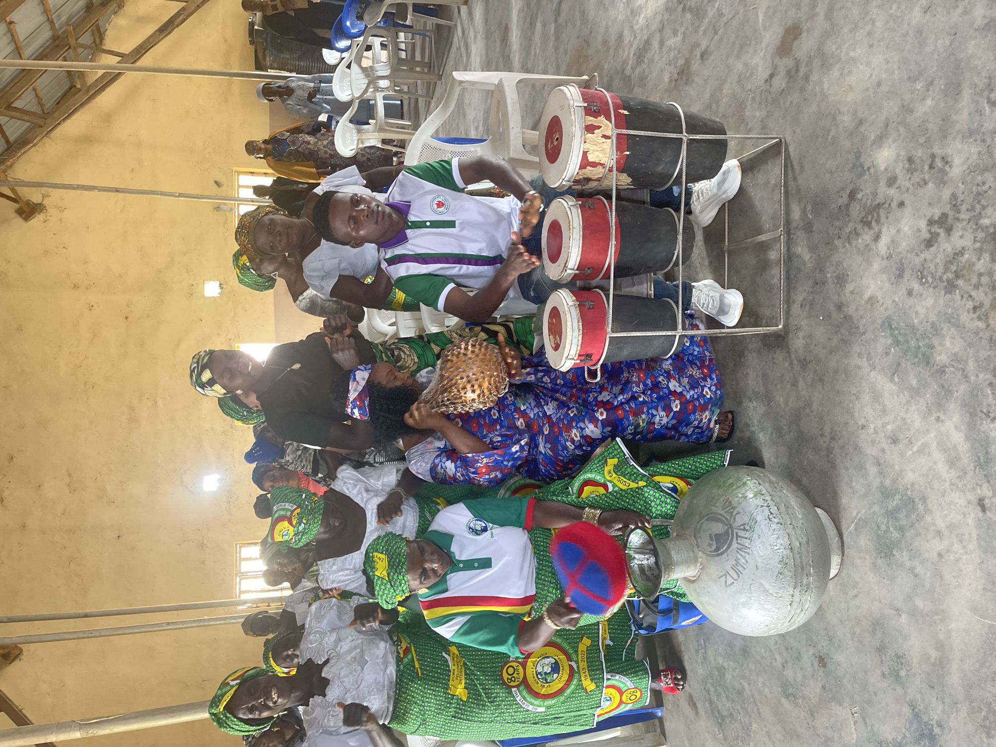 A group of people, predominantly women wearing vibrant traditional attire and headwraps, are gathered in an indoor space with a concrete floor and a wooden ceiling. The room appears to be a hall or a place of worship, with chairs arranged in rows and natural light filtering in from windows. In the foreground, a young man sits on a chair playing a set of three drums. To his left, a woman in a colorful dress holds a woven basket, and another woman in a green and white uniform is actively engaged with a large, metallic pot. Several other women, also in traditional clothing, are visible in the background, some dancing and others observing. The atmosphere suggests a celebration or a cultural event taking place in Mangu Town, Nigeria.