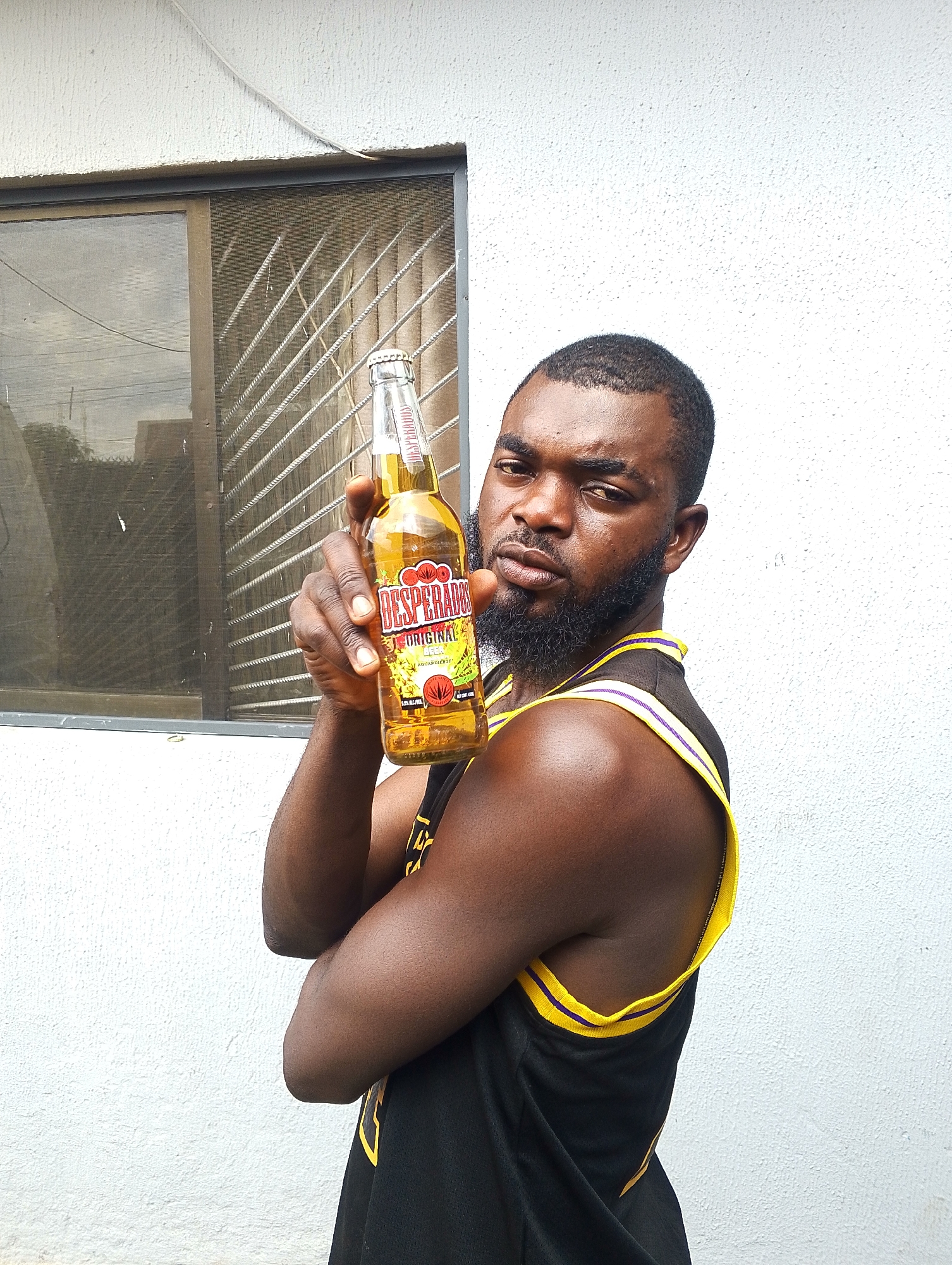 A man, with dark skin, a beard, and short hair, stands in front of a white, textured wall. He is wearing a black tank top with yellow and purple trim. He holds a bottle of Desperados Original beer in his right hand, which is raised. The bottle is made of clear glass and contains a golden-colored liquid. The label on the bottle is visible, featuring the brand name "DESPERADOS" in large red letters, along with text that appears to say "ORIGINAL" and "BEER." There is also a depiction of a cactus and some leaves on the label.
Behind the man, to his left, is a window with metal bars and a screen. The window reflects some of the environment, showing trees and a building in the distance. The sky appears overcast. The lighting suggests it might be daytime, possibly late afternoon. The man's expression is neutral, with a slight furrow in his brow. The scene appears to be outdoors, likely in a residential or urban area.
