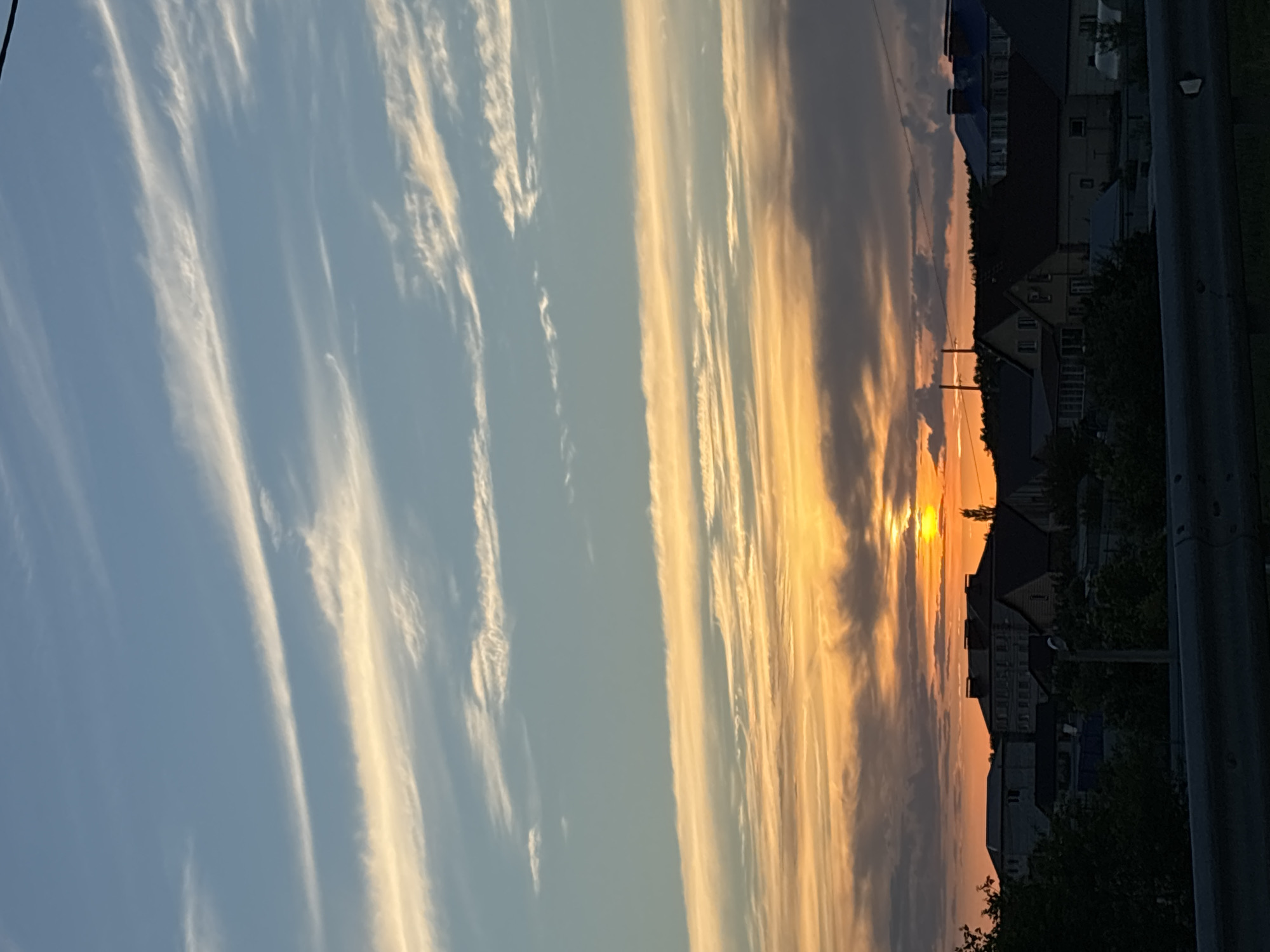 The image depicts a sunset over a semi-urban or suburban landscape. The sky is a gradient of blue at the top, transitioning to vibrant orange and yellow hues near the horizon, indicative of the setting sun. Wispy white clouds are scattered throughout the sky, catching the light from the sun. Below the sky, a line of buildings and houses is visible, silhouetted against the bright sky. These structures appear to be residential, with various roof shapes and sizes. Some greenery, likely trees and bushes, is present in the foreground and between the buildings, partially obscuring some of the structures. A grey metal barrier or guardrail is visible in the very bottom of the frame, suggesting the vantage point might be from a road or elevated walkway. The overall impression is a peaceful, late evening scene.