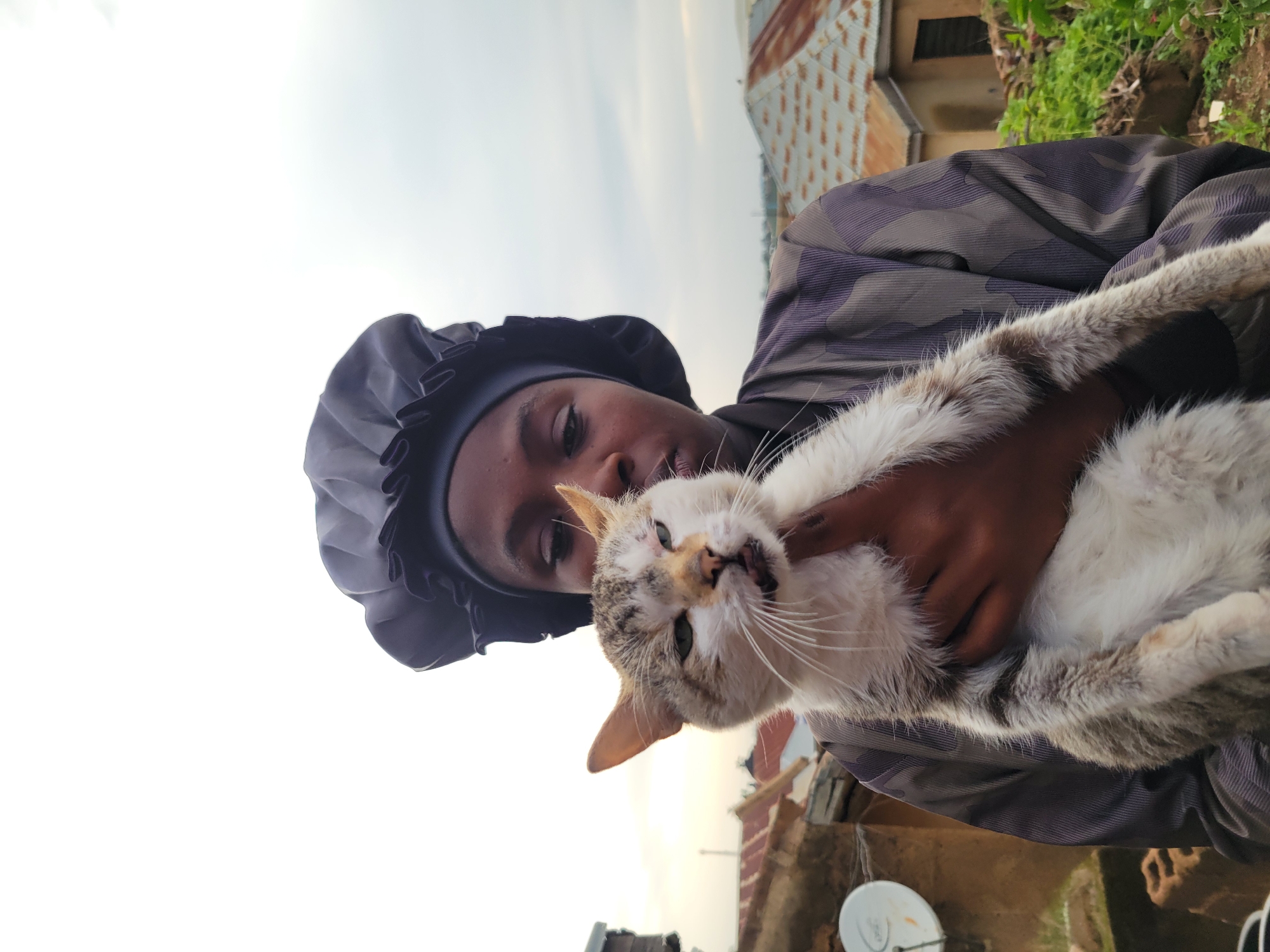 A medium shot captures a young person wearing a dark blue satin bonnet, holding a grey and white tabby cat. The person is dressed in a dark, patterned long-sleeved shirt with a camouflage-like design. The cat, held close to the person's chest, is looking towards the camera with its mouth slightly open. The background suggests an outdoor environment, possibly a rooftop or elevated area, with residential buildings and rooftops visible. The architecture of the buildings, with their corrugated metal roofs, indicates a setting in Nigeria, specifically within a district of Jos. The sky is overcast with a diffused light, suggesting either early morning or late afternoon. The overall impression is a casual, intimate moment between the person and the cat.