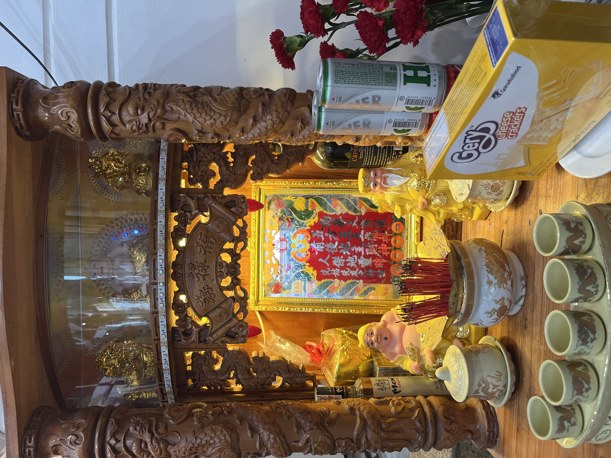 A detailed view of an ancestral altar, likely located in Hội An Ward, Vietnam. The altar features intricate, carved wooden posts and beams, potentially with dragon motifs. Within the altar, various items are arranged, including a framed image with red calligraphy, a golden statue of a deity (possibly the God of Prosperity), a small pink pig figurine, a jar filled with incense sticks, and a set of small cups arranged in a row. Food offerings are also present, consisting of a box of Gery cheese crackers and two cans of beer. Red carnations add a decorative touch. The altar appears to be illuminated by built-in lighting.