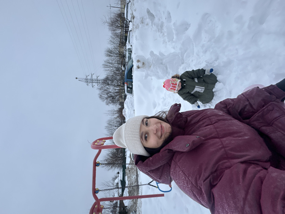 An outdoor, winter scene depicts a woman and a child enjoying the snow. The primary subject is a woman in the foreground, positioned on the right side of the frame, smiling warmly at the camera. She is wearing a light-colored, possibly off-white, knitted beanie and a dark reddish-purple puffer jacket. Her dark hair is visible beneath the hat.
Further back and slightly to the left, a small child is visible, partially obscured by the snow. The child is facing away from the camera, wearing a pink and white patterned knitted hat with ear flaps and a thick, dark green snowsuit or winter coat. They are sitting in a mound of snow, appearing to be engaged in play. Next to the child, a small, partially formed snowman is visible, indicating they might have been building it.
In the mid-ground, to the left of the woman, a red and yellow playground structure, possibly a swing set or climbing frame, is partially visible. The entire ground is covered in a thick blanket of white snow.
The background features bare trees, suggesting winter, and what appears to be a parking area with several vehicles, some of which are also covered in snow. A large metal utility pole or electrical pylon with power lines extends vertically towards the overcast, grey sky.
The overall impression is that of a casual, happy moment between an adult and a child during a snowy day, likely at a park or playground in an urban or semi-urban setting. There is no discernible text visible in the image.