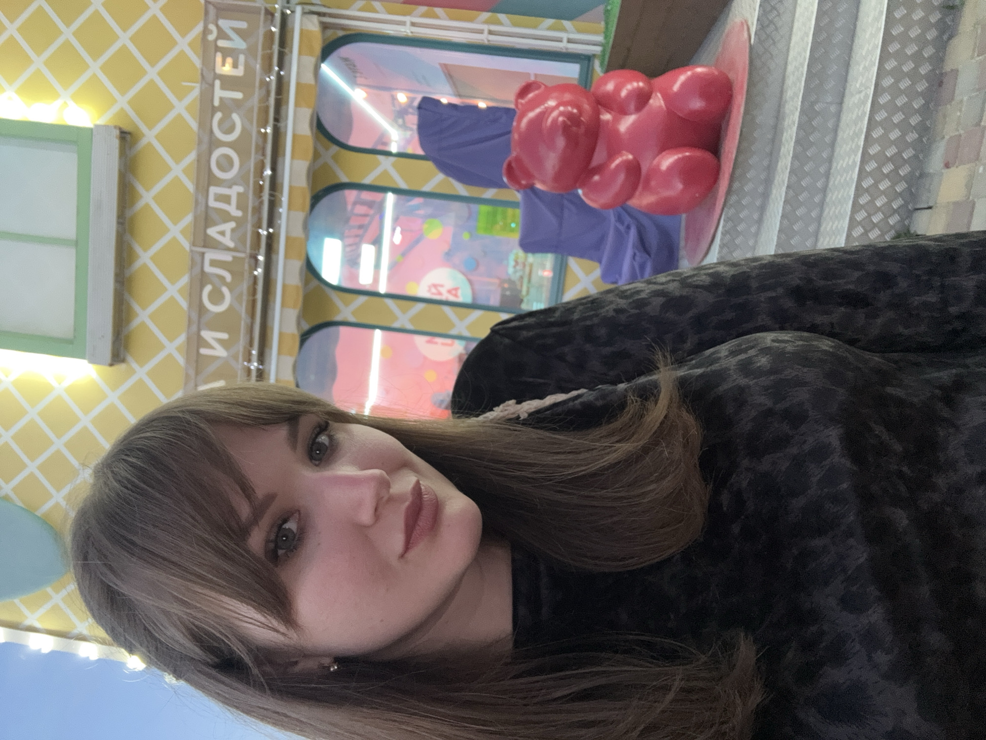 A medium shot shows a young woman with light brown hair and bangs, wearing a dark, patterned top, looking at the camera. Behind her, there is a building façade with yellow and white checkered pattern and the inscription "И СЛАДОСТЕЙ" which translates to "AND SWEETS" in Russian. There are also window displays, a pink teddy bear statue, and stairs, indicating a shop or attraction. The location is likely a sweet shop or candy store in Yevpatoriya, Ukraine. The time of day is likely daylight hours.