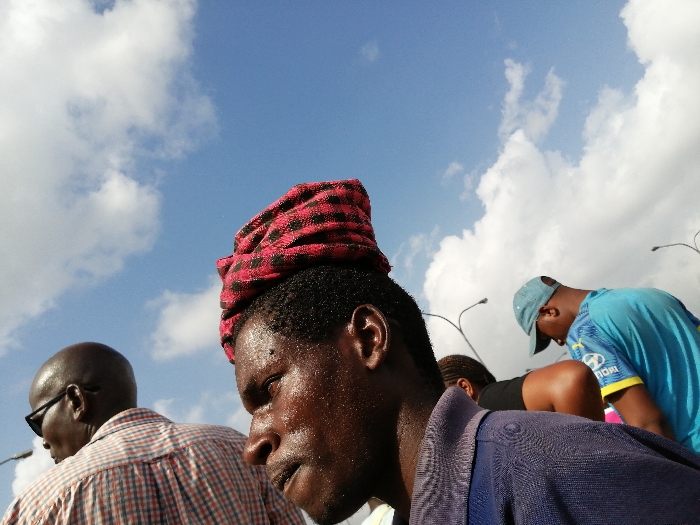 This outdoor shot, set against a bright blue sky dotted with scattered white clouds, captures a group of people in Dar es-Salaam, Tanzania. In the immediate foreground, a man with dark skin, his face glistening with sweat, is prominent on the right. He wears a dark blue shirt and a distinctive red and black checkered cloth wrapped around his head. His expression is serious and focused, as he looks downwards and to his right.
To the left and slightly behind him, another man is seen from the back, bald and wearing dark sunglasses and a light-colored plaid shirt. In the mid-ground on the right, a third individual, wearing a light blue t-shirt with yellow accents and a light blue baseball cap, is bent forward. A "Hyundai" logo is partially visible on their shirt. Another person's head and shoulder are visible behind the foreground man, also appearing to be bent or engaged in an activity.
The bright natural lighting indicates it is daytime, and the visible sweat suggests warm or hot weather. Several streetlights or utility poles are silhouetted against the sky in the background, hinting at a public or urban setting. The overall scene conveys a sense of earnest activity or observation within a busy environment.