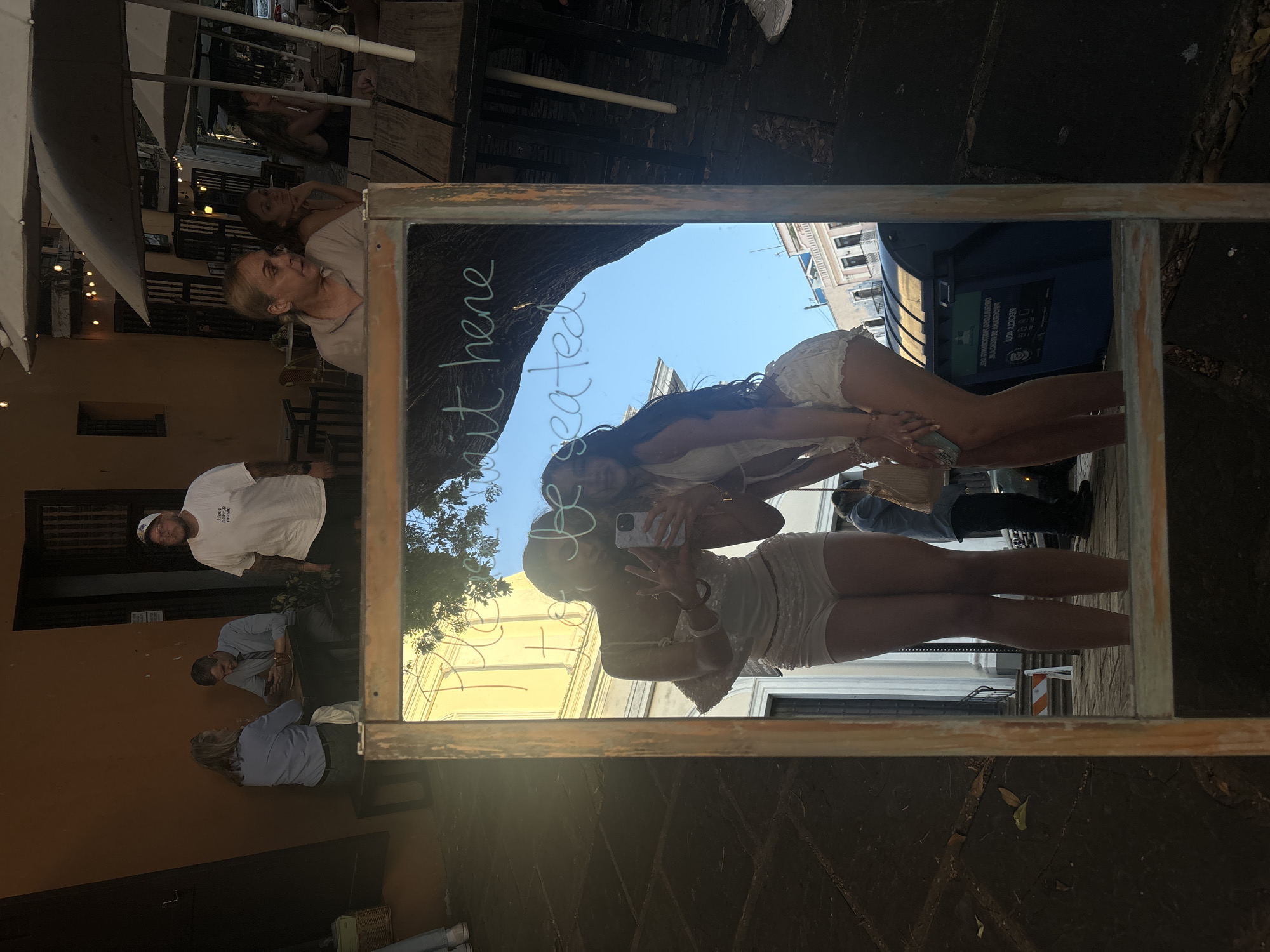 A medium shot captures a street scene reflected in a wooden-framed mirror. The reflection shows two young women in matching white outfits posing for a selfie, with one holding a phone. Behind them, a tree casts a shadow across the mirror's surface, partially obscuring the text "Please wait here to be seated" written in a light color. In the background of the reflection, several people are seated at outdoor tables. A man in a white t-shirt stands to the right, facing away from the mirror. The setting appears to be a bustling outdoor dining area in San Juan, Puerto Rico. The ground is paved with dark stones, and a blue recycling bin is visible in the reflection. The overall lighting suggests it is daytime.