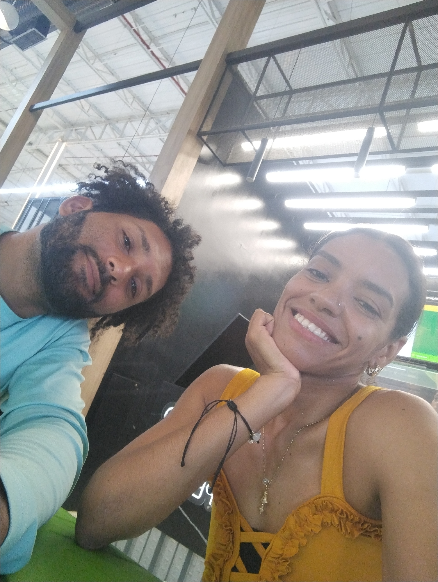 Two individuals are visible in a selfie-style photograph. The man, positioned on the left, has dark curly hair and a beard. He is wearing a light blue shirt. The woman on the right is wearing a yellow top and has dark hair pulled back from her face. She is smiling and resting her chin on her hand, which features a bracelet. A necklace is visible on her chest. The background reveals an interior space with an industrial-style ceiling that features metal framework and linear lighting fixtures. Based on the presence of the Sigo La Galeria shopping mall and its architectural style, the location is likely within this mall in the city of Porlamar, Venezuela. The couple is likely at a table within the mall.