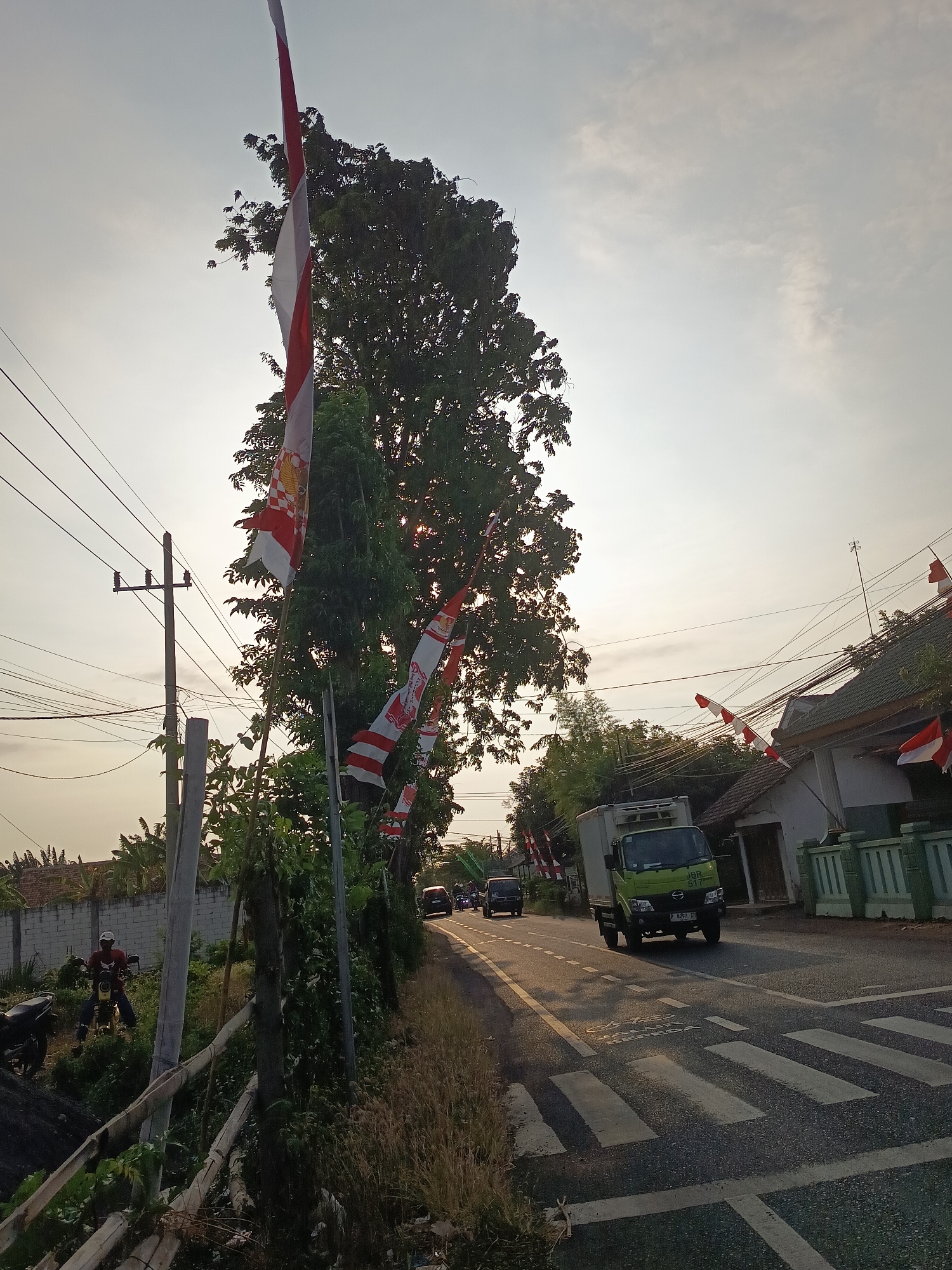 The image depicts a street scene in Kandangjati Wetan, Indonesia, bathed in the soft light of a setting sun. Several Indonesian flags, red and white, are prominently displayed, some hanging from tall trees and poles, others adorning the roofs of houses. These flags suggest a festive or patriotic occasion.
A road runs through the scene, with a green truck and other vehicles, including motorcycles, visible. One motorcycle, ridden by a person wearing a red shirt and a white cap, is parked on the side of the road near a wall. The road has a pedestrian crossing.
The environment includes lush green trees and vegetation lining the road. Electrical poles and wires are also present, indicating a developed area. The houses visible appear to be of modest construction. The sky is partly cloudy, with the sun low on the horizon, casting long shadows and creating a warm glow. The overall atmosphere is tranquil and rural.