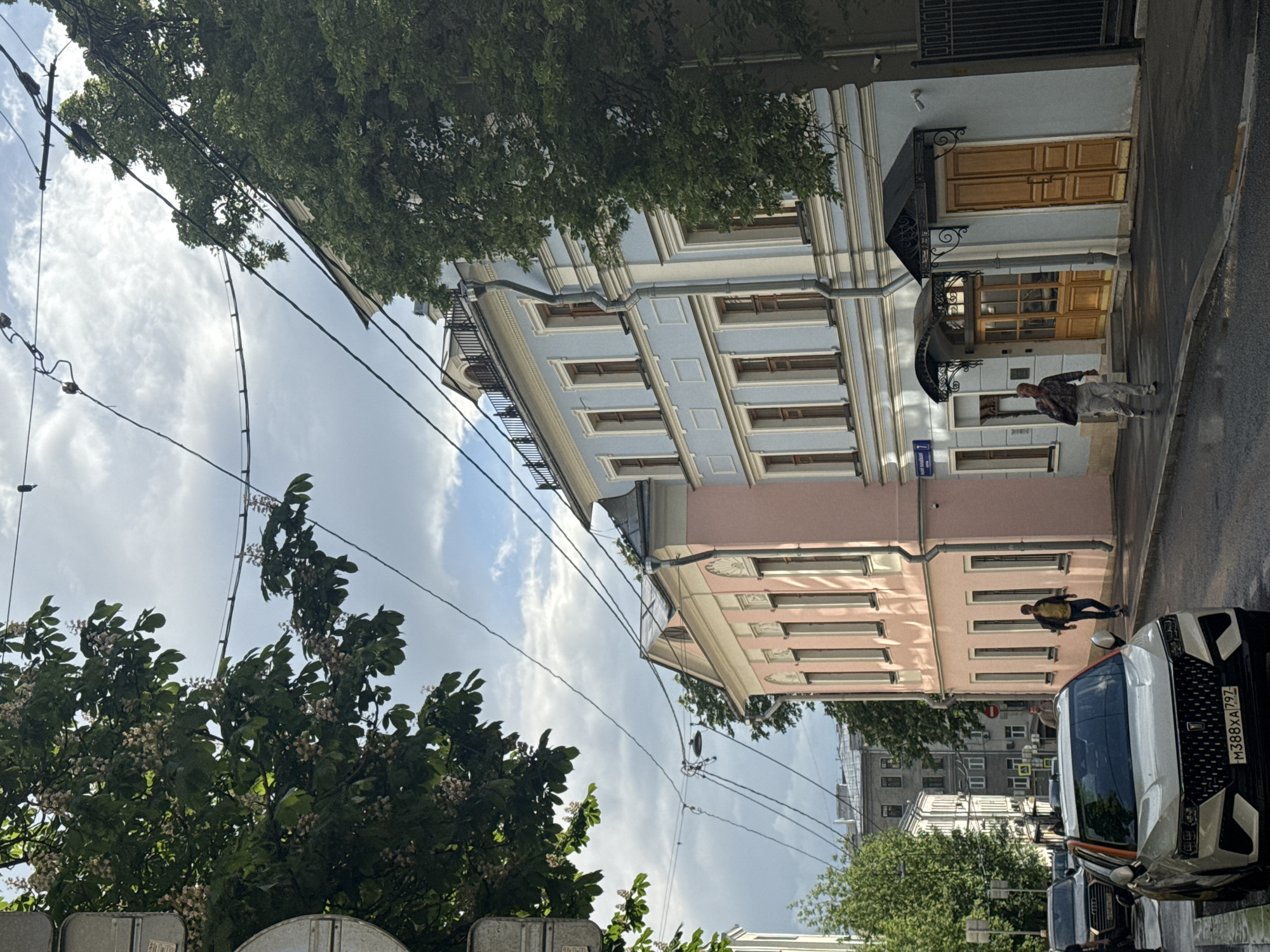 The image shows a street scene in Moscow, Russia. Two buildings, one painted light blue and the other pink, are visible on the right side of the frame. The street is wet, suggesting recent rain. Several people are walking on the sidewalk, and cars are parked along the street. The trees with white blossoms indicate it is likely springtime. Power lines traverse the scene against a cloudy sky. A building address sign is visible on the facade of the light blue building. The license plate M388XA797 is visible on a vehicle on the street.