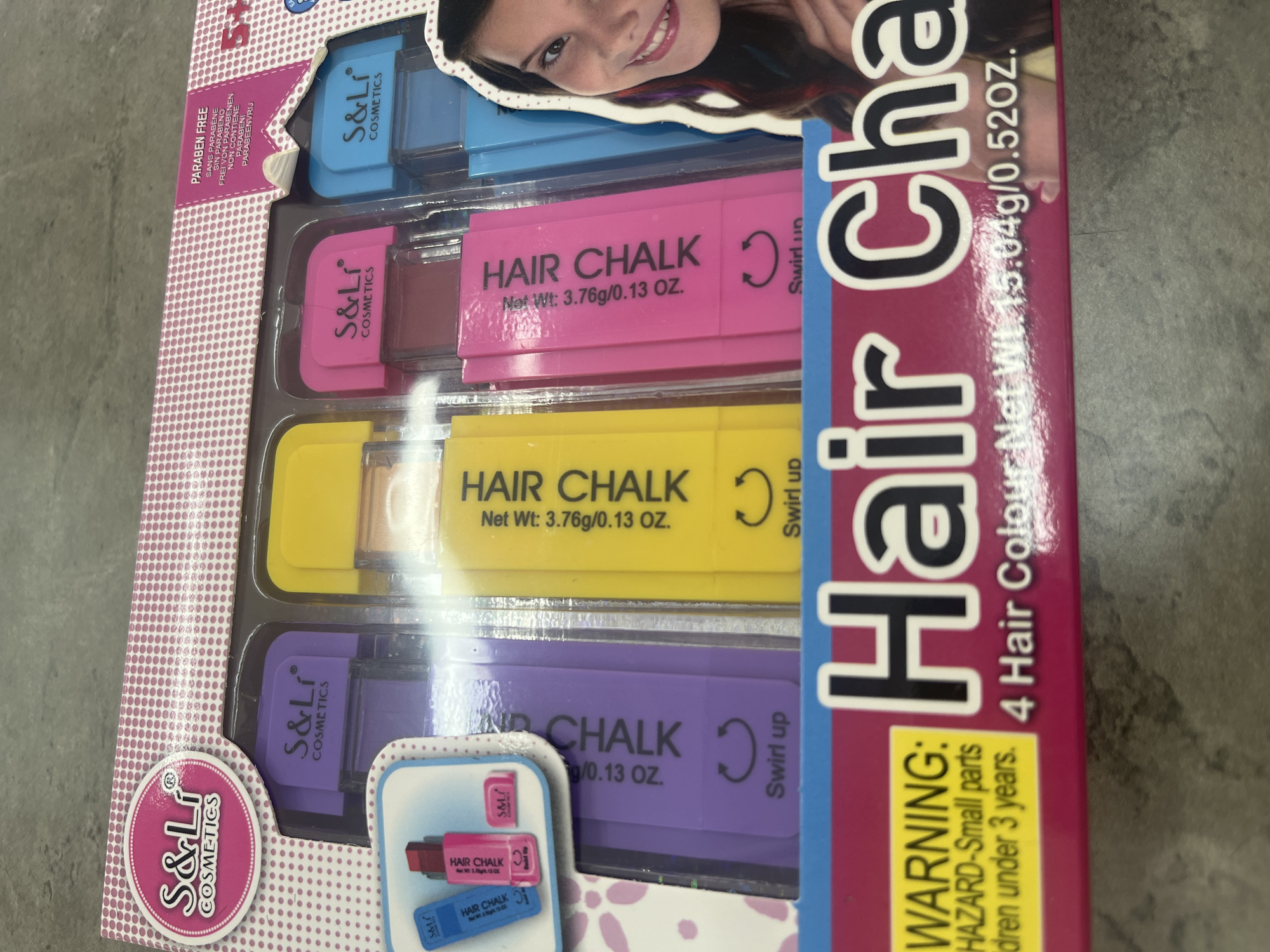This is a close-up, eye-level, photograph of a boxed set of S&Li Cosmetics Hair Chalk. The box is primarily pink and white with polka dots and has a large illustration of a girl with colorful streaks in her hair on the right side. The box displays four individual hair chalk applicators in purple, yellow, pink, and blue. Each applicator has the brand name "S&Li COSMETICS" printed on it. Text on the applicators reads "HAIR CHALK Net Wt: 3.76g/0.13 OZ." and "Swirl up". A "WARNING: HAZARD-Small parts Children under 3 years." label is visible on the lower left of the box. The product name "Hair Cha" is printed largely across the bottom of the box. The background appears to be a textured, gray surface, possibly a floor or table. There is no discernible information that would place this scene in the city of Uzhhorod, Ukraine.