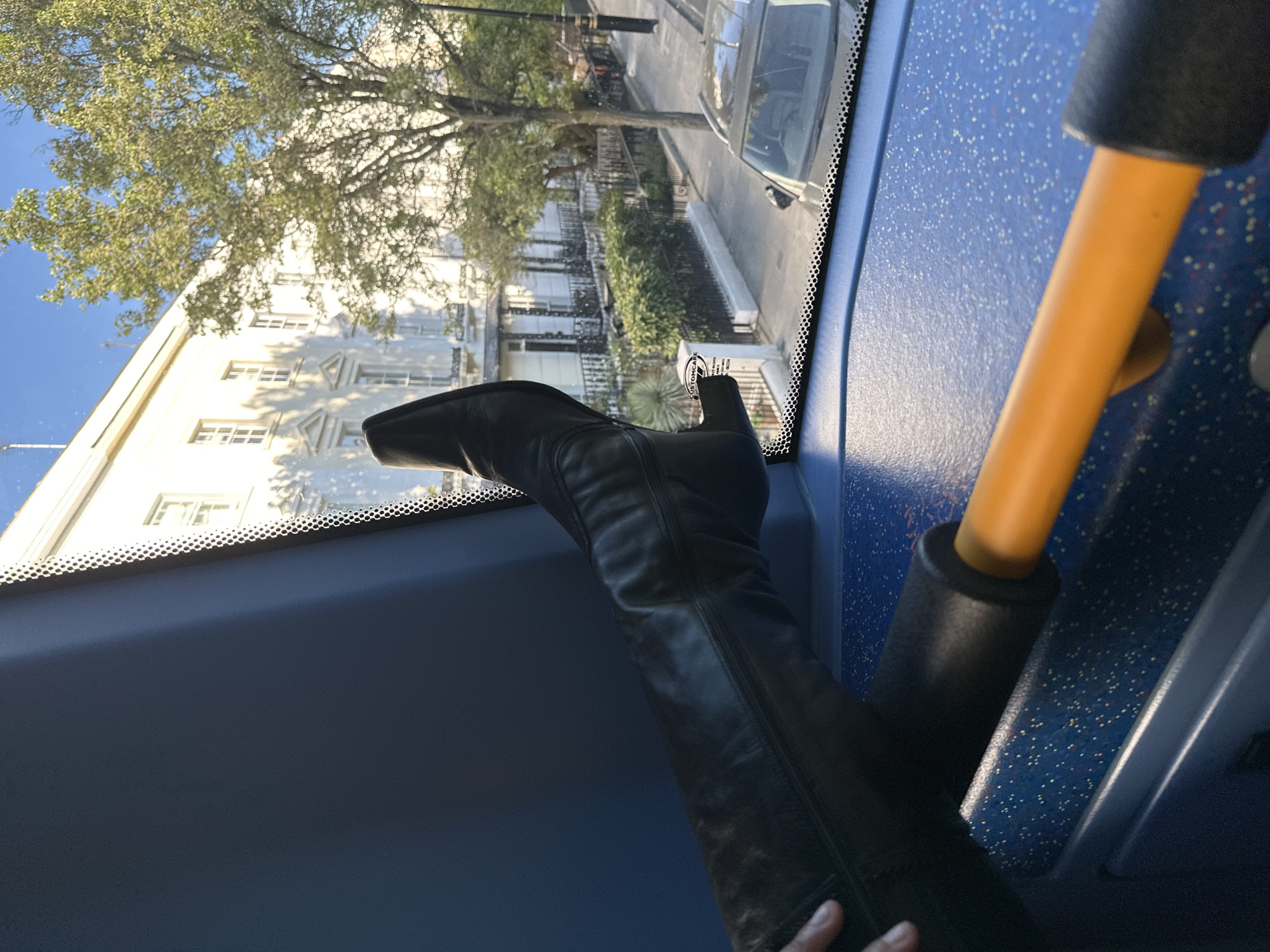 The image captures a first-person perspective from inside a vehicle, likely a bus given the interior features and elevated view, looking out onto a sunny street scene. The composition is tilted counter-clockwise.
In the immediate foreground, a person's left leg, clad in a long, black leather boot, is prominently featured, resting casually on what appears to be the dashboard or a front ledge of the vehicle. A hand is visible near the bottom edge of the frame, lightly touching the boot. The interior of the vehicle is characterized by a blue surface, possibly a floor or lower wall, speckled with white, red, and yellow dots. An orange handrail with a black grip is visible on the right side, further suggesting a public transport vehicle. The clear front window (windscreen) features a black dotted frit band along its top edge.
Through the window, a bright urban street unfolds. Across the street, a row of elegant, light-colored terraced houses, typical of London architecture, are visible with multiple windows and some decorative architectural elements, including triangular pediments. Lush green trees with dense foliage partially obscure the buildings and line the street. A paved road is visible with at least one parked car, and black wrought-iron railings separate the street from the house-fronts. The sky above is a clear, bright blue, indicating sunny weather.
The time of day appears to be bright daytime, with ample sunlight illuminating the buildings and casting shadows from the trees. The relaxed posture of the leg implies a casual or comfortable journey. A small sticker on the windshield, near the toe of the boot, has partially legible text, including "CUSTOMCAR" or "CUSTOMCARE". The architectural style and street layout strongly suggest a London setting, aligning with the provided context.