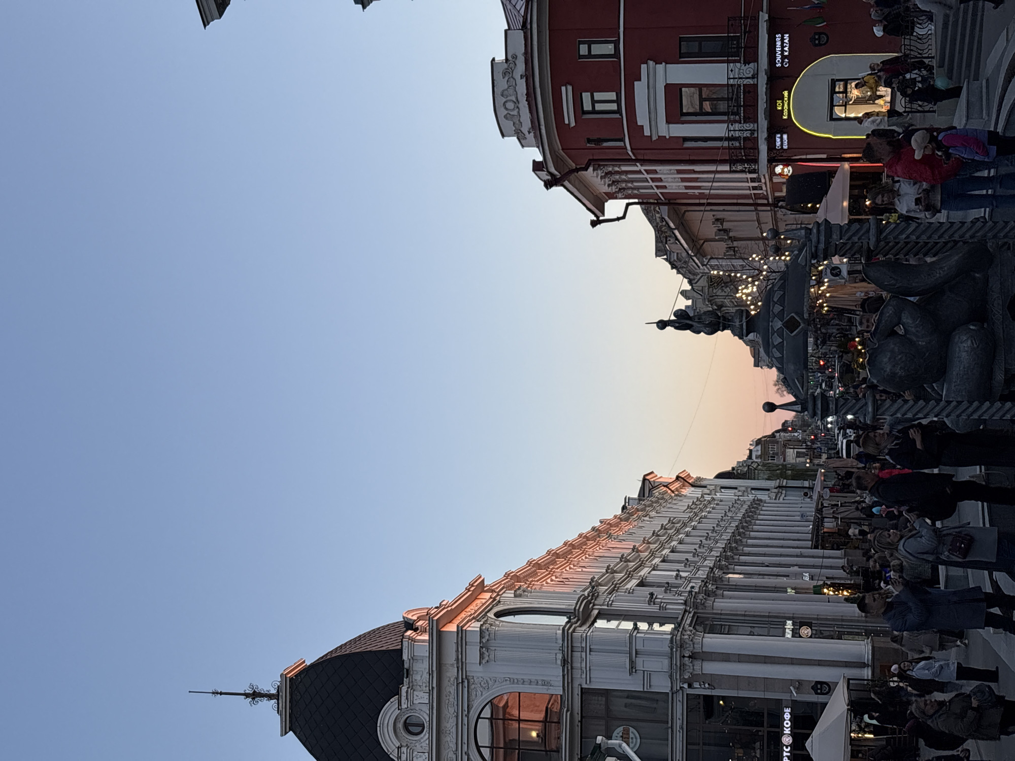 The image depicts a street scene in Kazan, Russia at dusk. Two buildings frame the view of the sky, with one building white and ornate with numerous columns and the other red with white trim. Between them is a pedestrian zone crowded with people. A statue sits centrally in the shot. The buildings have businesses at ground level, while the sky shifts from blue at the top to orange at the horizon.