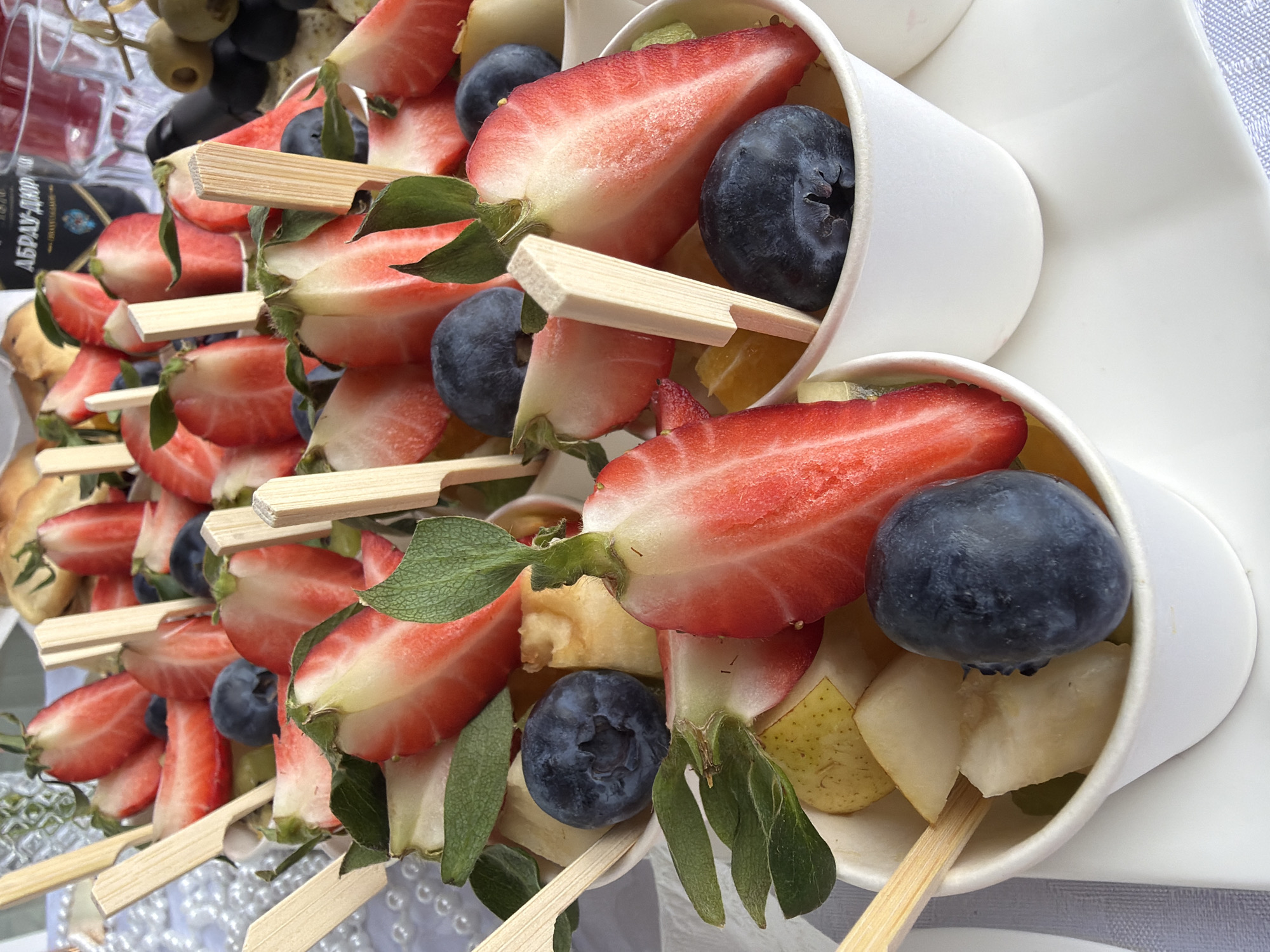This image presents a close-up view of an array of fresh fruit skewers, thoughtfully arranged in small, individual white paper cups. Each cup contains a wooden skewer holding a selection of fruits, predominantly halved strawberries and whole blueberries. Accompanying these are chunks of lighter-colored fruits, identifiable as pear, and possibly orange or apple, adding variety to the mix. Many of the halved strawberries are presented with their vibrant green leaves still attached, enhancing the visual freshness and presentation. The skewers themselves are flat, made of light-colored wood, and feature a wider, paddle-like end designed for easy handling.
The white paper cups are tilted and densely packed together, suggesting a buffet or catering setup for an event. They rest on a light-colored surface, possibly a subtly patterned tablecloth. In the softly blurred background, towards the upper left, an arrangement of grapes, including dark and olive-green varieties, can be glimpsed, indicating other fresh produce is part of the spread. To the left and slightly further back, a dark bottle or container is visible, featuring a label with distinct Cyrillic text.
The visible text on the bottle's label clearly reads "АБРАУ-ДЮРСО" (ABRAU-DURSO). This is a well-known Russian brand, primarily associated with sparkling wines, which provides a strong contextual cue aligning with the specified location of Chaikovsky, Russia. The bright and even lighting across the scene suggests a well-lit indoor environment or an outdoor event under clear conditions. The overall impression is one of a health-conscious and visually appealing refreshment option served at a social gathering or celebration.