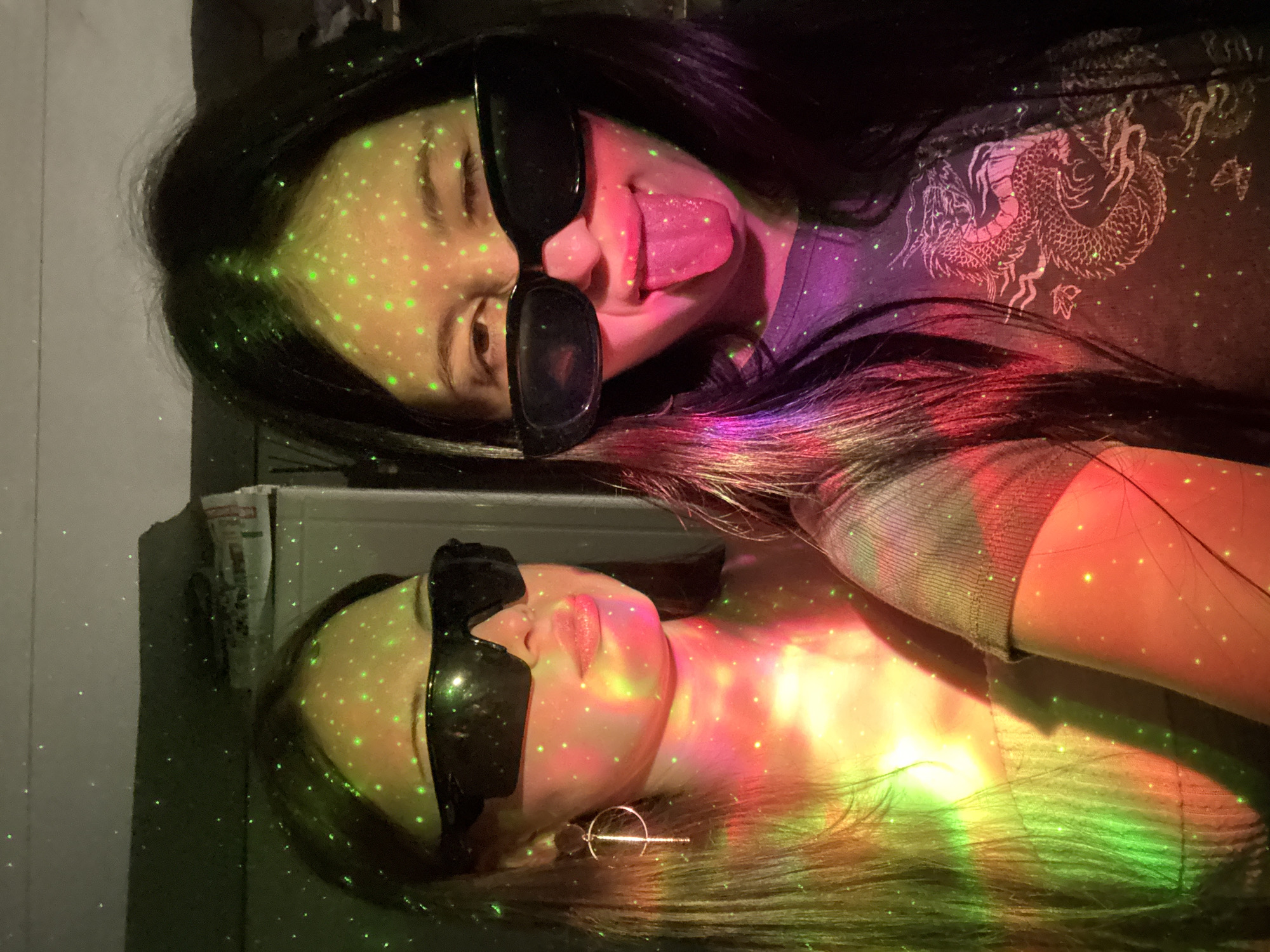 This media file captures two individuals in a dimly lit indoor setting, illuminated by vibrant, multicolored laser lights. The image is oriented vertically, suggesting the subjects are lying down, or the photo was taken from an overhead perspective and rotated.
The main subjects are two young individuals, likely women, both with long, dark hair. They are wearing dark sunglasses. The person in the foreground (top of the image) wears a dark t-shirt featuring a prominent white or light-colored dragon design on the shoulder/back. This person is playfully sticking their tongue out, showing a relaxed and expressive demeanor. Green laser dots are scattered across their face and hair, and their skin is also tinged with red and purple light.
The second person, positioned behind and slightly below the first, also wears sunglasses and has long dark hair. This individual appears calmer, with a serene expression. Their face, neck, and hair are covered in a mix of green laser dots and broad washes of red, yellow, and green light, creating a rainbow effect. A metallic earring is visible on their right ear. The fabric of their clothing appears lighter in color than the first person's, and the texture of their sleeve is visible.
The activity appears to be a casual photoshoot or a moment captured during an event with special lighting, possibly a party or concert, given the disco-like laser projections. The time of day is certainly night or a very dark room, as indicated by the reliance on artificial lighting. There are no clear geographical location cues within the image itself, beyond an indoor environment. No text is visible in the image. The overall mood is playful and energetic due to the vibrant lighting and expressive poses.