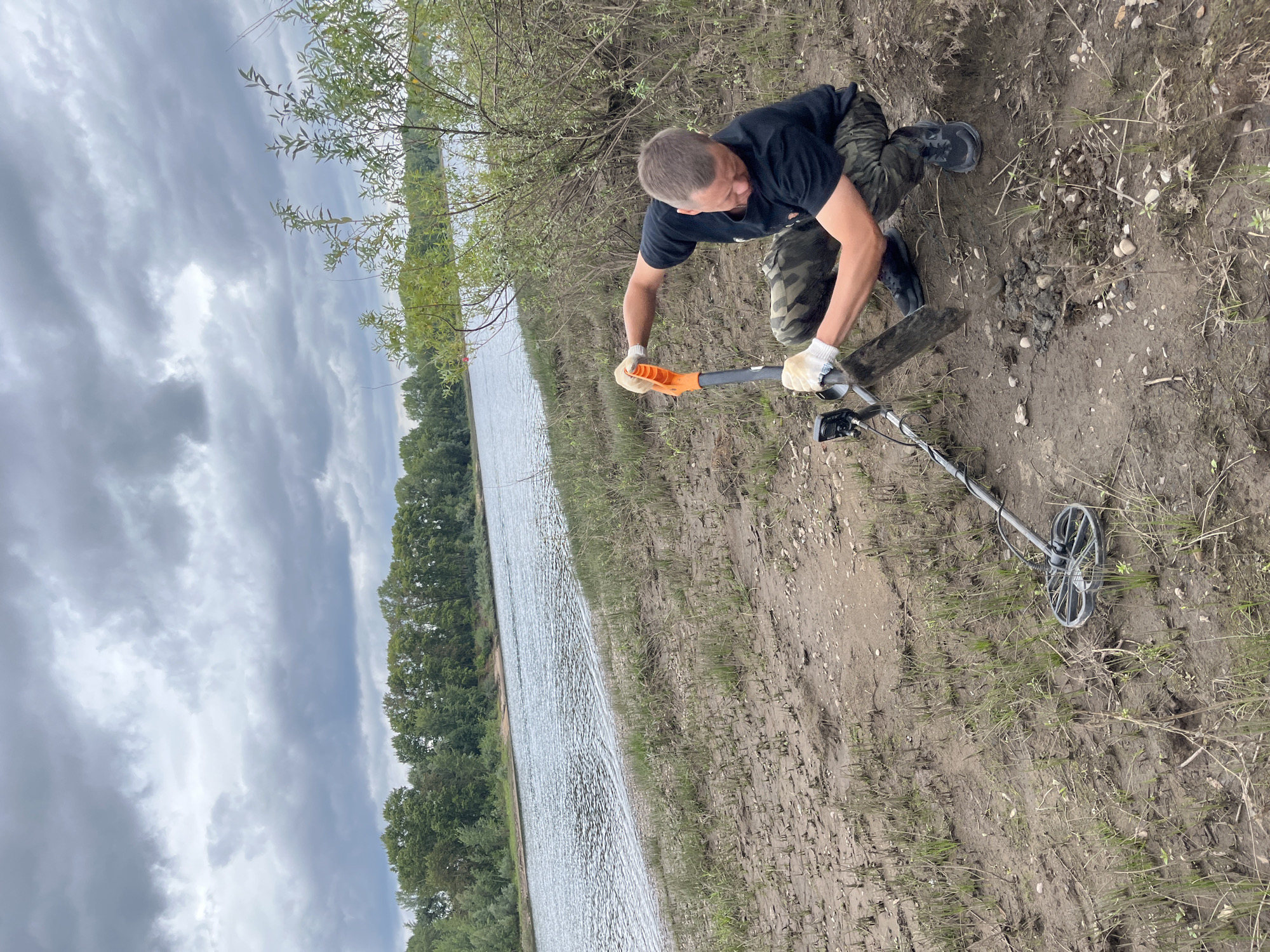 A man wearing a black t-shirt, camouflage pants, white gloves, and black shoes is metal detecting on the muddy bank of a wide, slow-moving river under a cloudy sky. He is squatting and using an orange-handled shovel to dig a small hole in the dirt. A metal detector with a circular search coil is visible in the foreground. The background shows a dense line of green trees on the opposite bank of the river, and a few scattered bushes on the near bank. The scene appears to be in an undeveloped area, possibly a park or natural reserve, not within the city of Chaikovsky, Russia itself, but rather in its surrounding natural landscape.