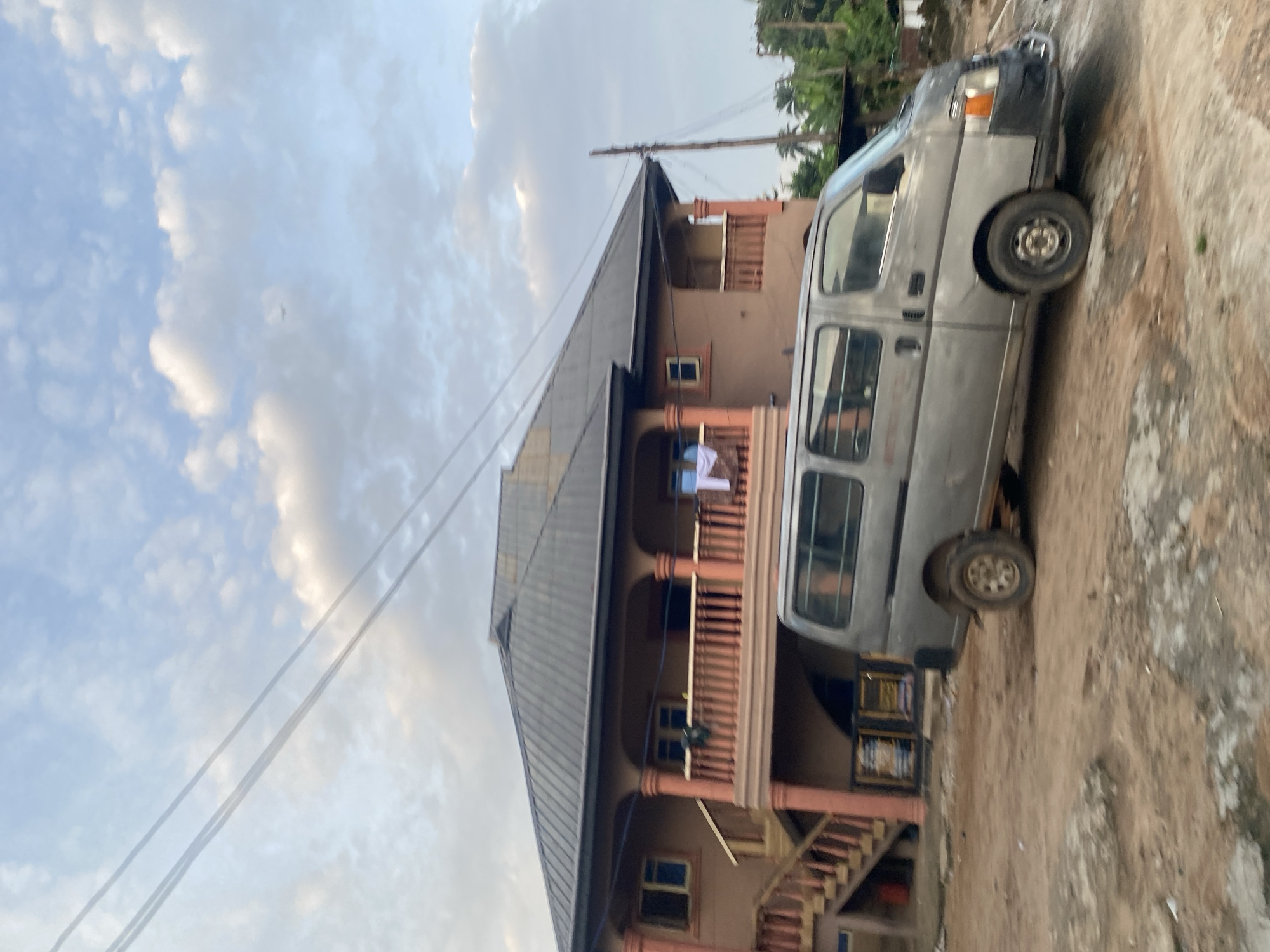 The image captures a static street scene in Enwan, Nigeria, under a partly cloudy, light blue sky traversed by two parallel power lines. A small, indistinct object, possibly an airplane, is visible in the upper right sky. The primary subject is a two-story building with a dark, possibly corrugated metal, gabled roof and reddish-brown walls, featuring multiple windows and two second-story balconies with reddish-orange railings and pillars. An exterior staircase, matching the balcony railings, ascends to the second floor on the left. A piece of white laundry hangs over the railing of the left balcony. In the foreground, a worn, light grey van, possibly a panel van, is situated on an unpaved, reddish-brown dirt road, partially obscuring the lower section of the building. The van's wheels are visibly dirty, and its front bumper appears damaged or discolored. To the right of the building, a rustic wooden utility pole stands amidst green tropical foliage, including palm trees. No individuals are visible in the scene.