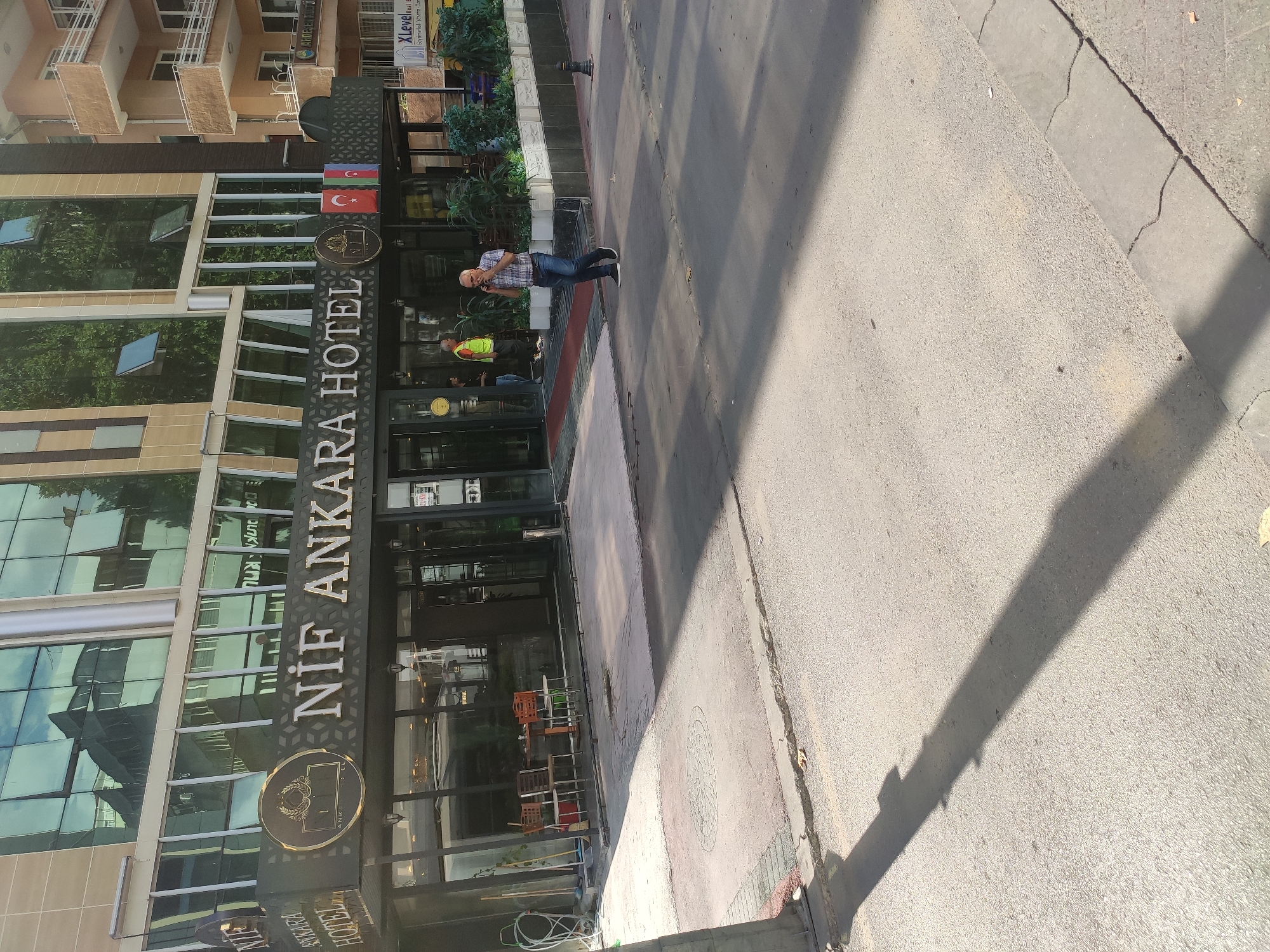 The image shows the exterior of the NIF Ankara Hotel in Ankara, Turkey. The hotel has a modern facade with large windows. A man in a plaid shirt and jeans is walking on the sidewalk in front of the hotel, talking on his phone. Another man in a high-visibility vest is standing near the entrance of the hotel. There are potted plants and tables and chairs visible on the sidewalk in front of the hotel. The street in front of the hotel is paved and has a sidewalk. Shadows are cast across the street and sidewalk from the buildings and a pole.