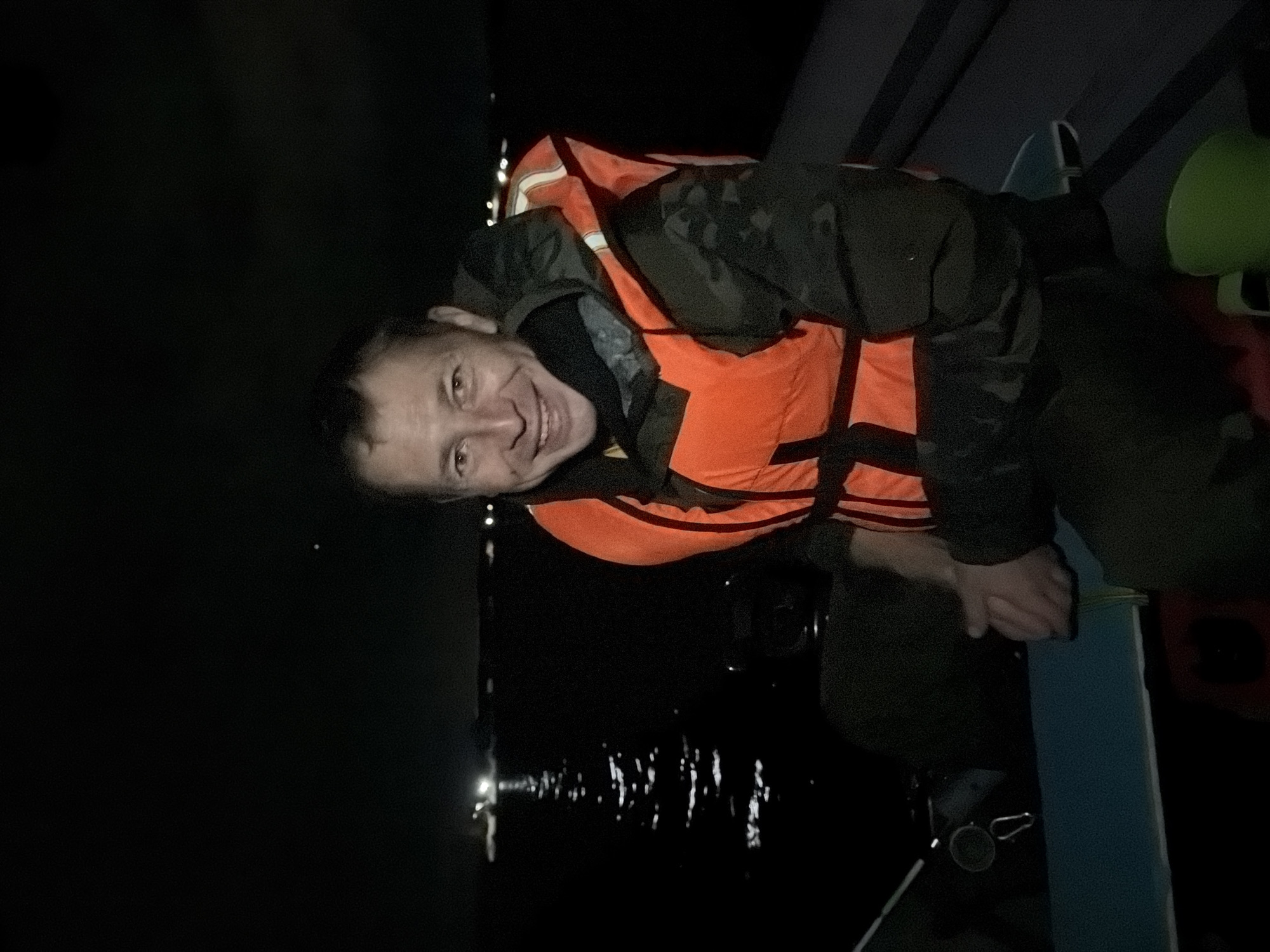 A man wearing an orange life vest and a dark, camouflage-patterned jacket is seated in a boat at night. He is smiling and looking at the camera. The boat appears to be on a body of water, with lights visible in the distance, suggesting it is near a populated area or a port. The water reflects the lights, creating a shimmering effect. The background is dark, indicating it is nighttime. The man's hands are clasped in front of him. The scene is consistent with activities like night fishing or boating in or around the city of Chaikovsky, Russia, given its location on the Kama River.
