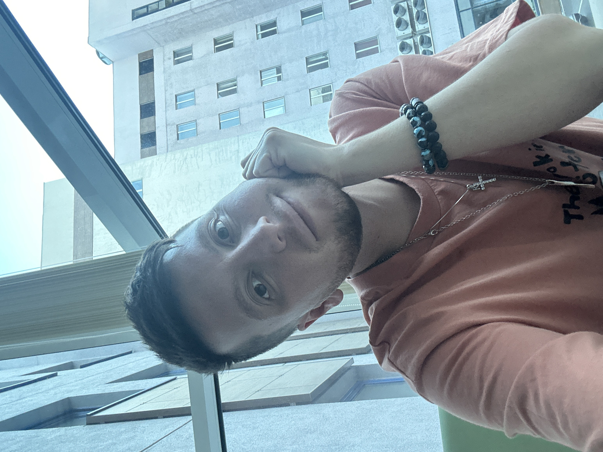 A selfie of a young man with short, dark hair and a beard is taken from the chest up. He is wearing an orange t-shirt and has two necklaces on, one with a cross pendant and another with a cylindrical pendant. He is also wearing two beaded bracelets on his right wrist. His right hand is resting on his cheek, and he is looking at the camera with a slight smile. In the background, there is a modern building with many windows. The man is positioned in front of a large window, with a view of the building and a clear sky. The building appears to be a residential or office building, common in urban areas. The lighting suggests it is daytime, possibly late afternoon, given the angle of the light.