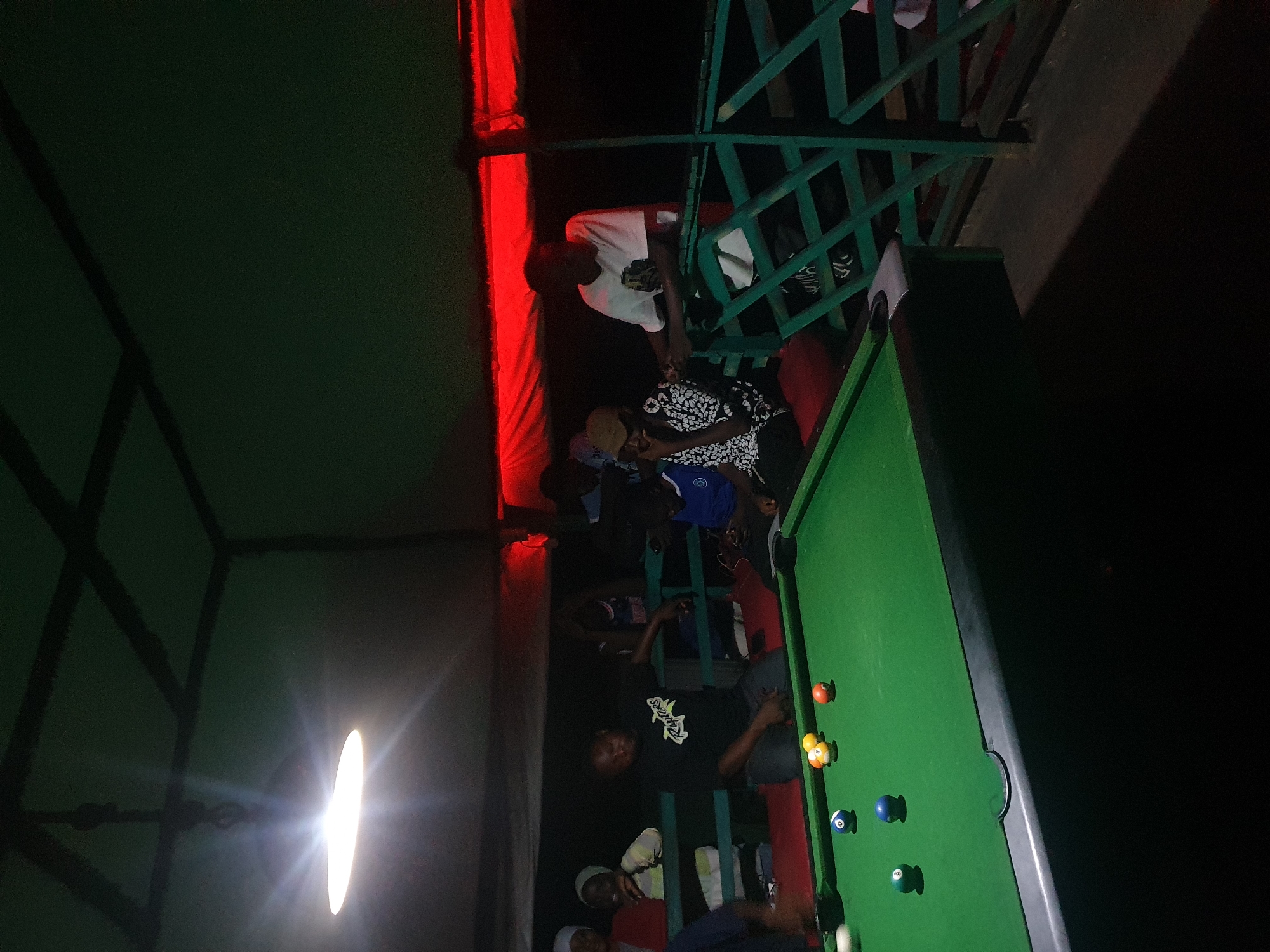 The image depicts an indoor billiard hall in Lokoja, Nigeria, illuminated by a bright overhead lamp and the red glow of lighting above. In the foreground, a green billiard table is set up with several balls scattered across its surface, including a blue ball, a solid yellow ball, a striped yellow ball, and a solid orange ball. In the middle ground, a group of people are gathered around the table. A man in a black t-shirt with the word "Raptors" printed on it is kneeling by the table. To his right, another man in a white t-shirt stands near a green railing. Further back, other individuals can be seen standing and sitting, some wearing head coverings. The environment appears to be somewhat dimly lit, with a textured ceiling structure visible above. The scene suggests a casual gathering, possibly a social event or a casual game of billiards.