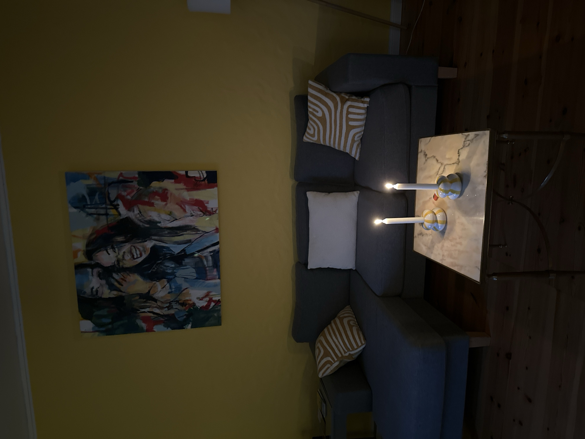 In Ammeldingen bei Neuerburg, Germany, a cozy living room is dimly lit by two lit candles on a marble-topped coffee table. The coffee table has a gold-toned metal frame. Behind the coffee table, a grey sectional sofa is arranged with two throw pillows: one white and one with a yellow and white abstract pattern. A large, colorful abstract painting depicting two laughing women hangs on a yellow wall above the sofa. The floor is made of warm-toned wooden planks.