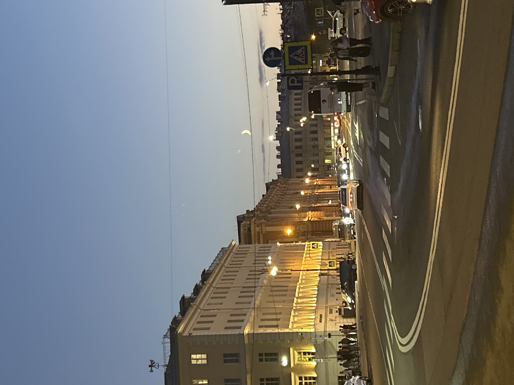 An evening street scene in an urban environment shows a crescent moon visible in the twilight sky above tall, ornate buildings with illuminated windows. The street is paved and features white lane markings and a crosswalk. Several cars are on the road, some in motion and some parked. Pedestrians are visible on the sidewalks, some walking and some standing. Traffic signs are present, including one indicating parking and another a pedestrian crossing. The overall impression is of a city preparing for night.