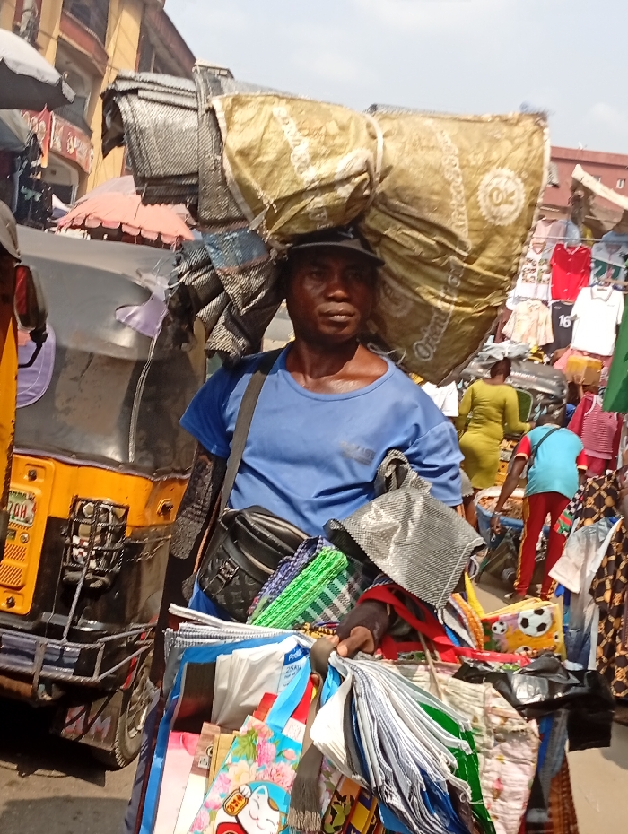 The image captures a bustling street scene, likely a market, during daylight hours under bright, sunny conditions. The central figure is a man in the foreground, facing forward and looking directly at the viewer. He wears a blue t-shirt and a dark baseball cap.
He is carrying a significant load on his head, consisting of at least two large, possibly woven, sacks or bundles. One prominent bundle, goldish-yellow in color, features partially visible text that includes "Orient" and a circular logo. Another bundle behind it is darker. He also wears a black fanny pack or waist bag strapped across his chest. In his hands, he carries a substantial display of various folded items, likely bags or fabrics, indicating he is a vendor. These items are diverse in color and pattern, including green checkered designs, plain white, blue, and brightly colored floral patterns. Some specific designs include a white bag with "25 kg" and "Pro" visible, another with a cat-like character (resembling a Maneki-neko), and one with a soccer ball pattern.
In the background, the environment is busy. To the left, part of a yellow and black auto-rickshaw (tuk-tuk) is visible, with "7DH" printed on a plate. Further back, buildings and other market stalls with goods are discernible. To the right, various items of clothing, such as red and white t-shirts, are hung on display, suggesting a clothes market. Several other people are present in the background, some partially visible and blurred, such as an individual in a lime green dress and another in a blue top and red pants. The man's expression is neutral, perhaps showing a degree of concentration or the effort of carrying his wares. The overall impression is one of active commerce in a vibrant urban setting.