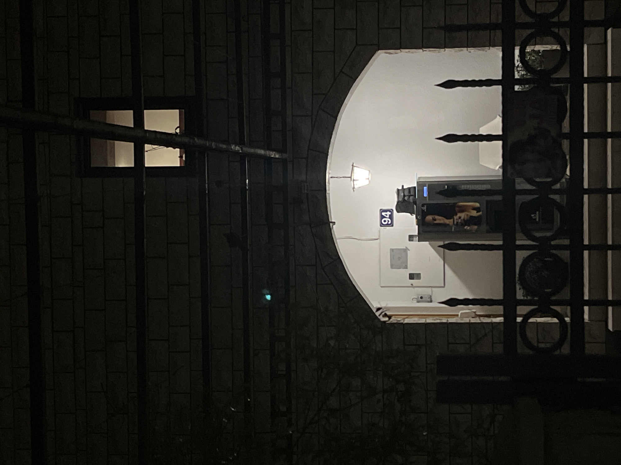 At night, a street in Budva, Montenegro, reveals a building facade with a brick pattern. A small window emits light, while an arched doorway frames a brightly lit interior. Inside, a number "94" is visible, along with a modern appliance. Outside, an ornate wrought-iron gate partially obscures the view. The scene suggests a residential or commercial property within the urban environment.