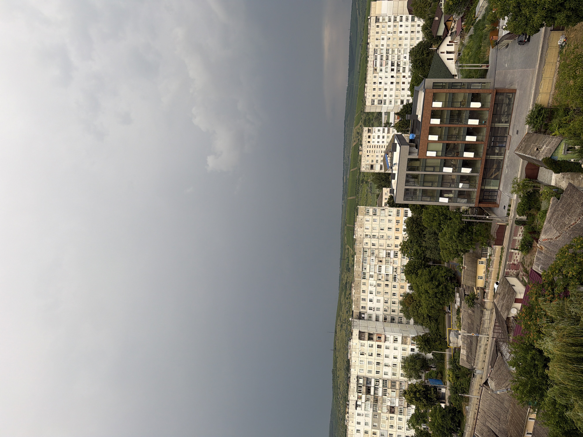 The image depicts an aerial view of a section of the city of Strășeni, Moldova, characterized by a mix of urban development and greenery. Dominating the upper portion of the frame is a vast, dark, and cloudy sky, suggesting an impending storm or overcast weather. Below the sky, a broad, undulating landscape stretches across the horizon, appearing as green fields or vineyards, with some evidence of terracing.
In the midground, several multi-story apartment buildings are visible. These buildings are predominantly white or light-colored, with numerous windows and balconies, indicative of Soviet-era architecture. Trees surround these buildings, creating a dense green canopy. A more modern, glass-fronted building with a brown facade stands out amongst the older structures, featuring large windows that reflect the sky. Adjacent to this modern building, a smaller white house with a dark roof is situated on a grassy slope.
The foreground reveals a more densely populated residential area with smaller houses, many featuring grey, sloped roofs. Walls and fences delineate property boundaries, and the area is interspersed with trees and vegetation. A dark-colored car is parked on a paved area near the modern building. The overall impression is one of a city transitioning between older and newer architectural styles, set against a backdrop of agricultural land and a dramatic sky.