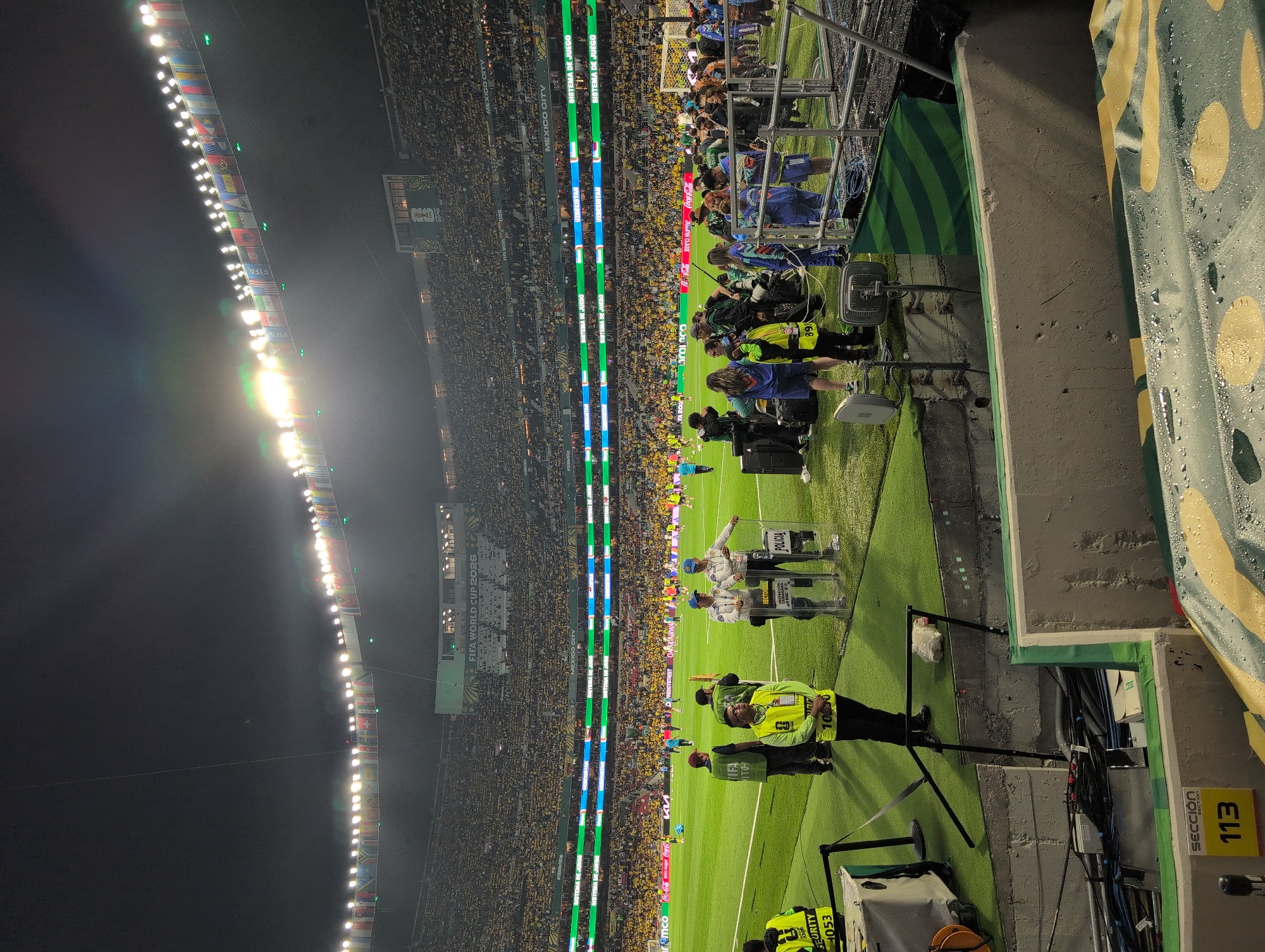A nighttime aerial shot of a packed stadium in Mexico City, Mexico, shows a large crowd of spectators wearing yellow and green, suggesting support for the Mexican national team. The stadium is illuminated by bright floodlights, and flags of various nations are displayed along the upper rim of the stadium. In the foreground, several security personnel and police officers are visible on the field, some holding transparent shields. The field itself is green turf, and there are various pieces of equipment and cables scattered around the perimeter. The overall impression is of a well-attended sporting event, likely a soccer match, in a major urban center.